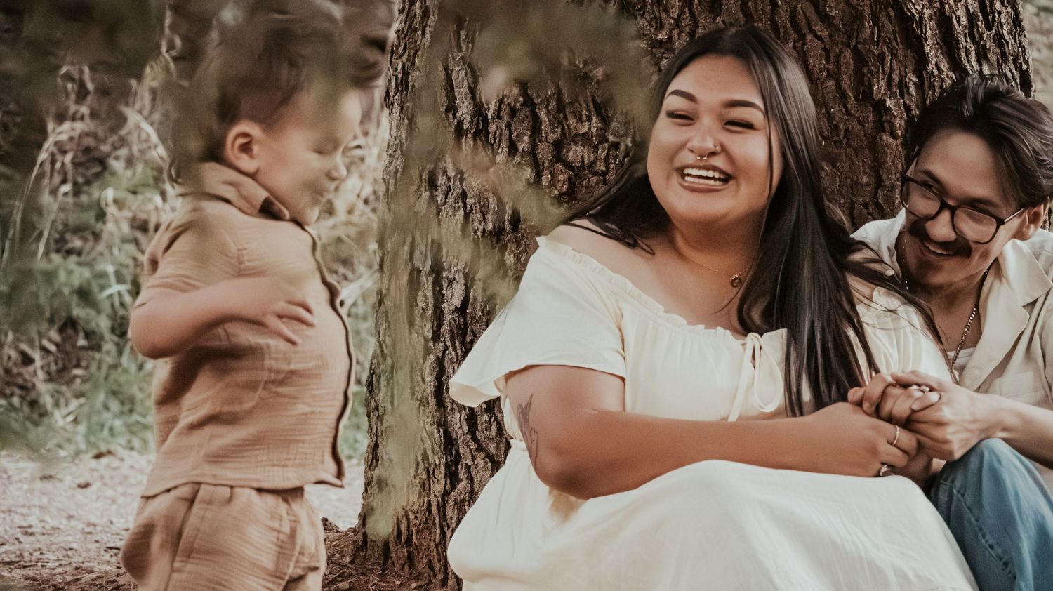 Family laughing together outdoors near a large tree, mother in white dress, father and young boy in tan outfit.