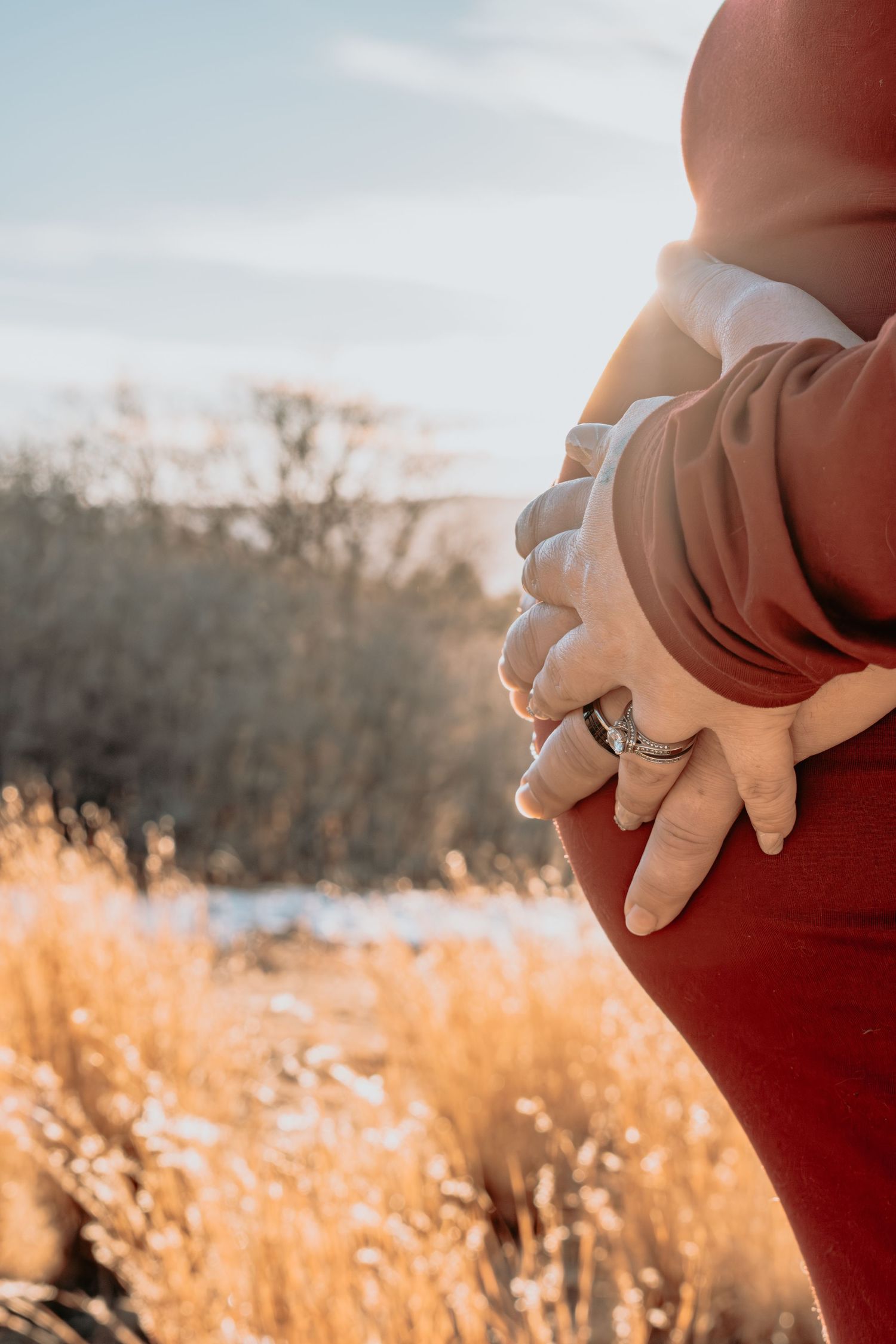 Close-up of couple holding pregnant belly in red dress with golden field background.