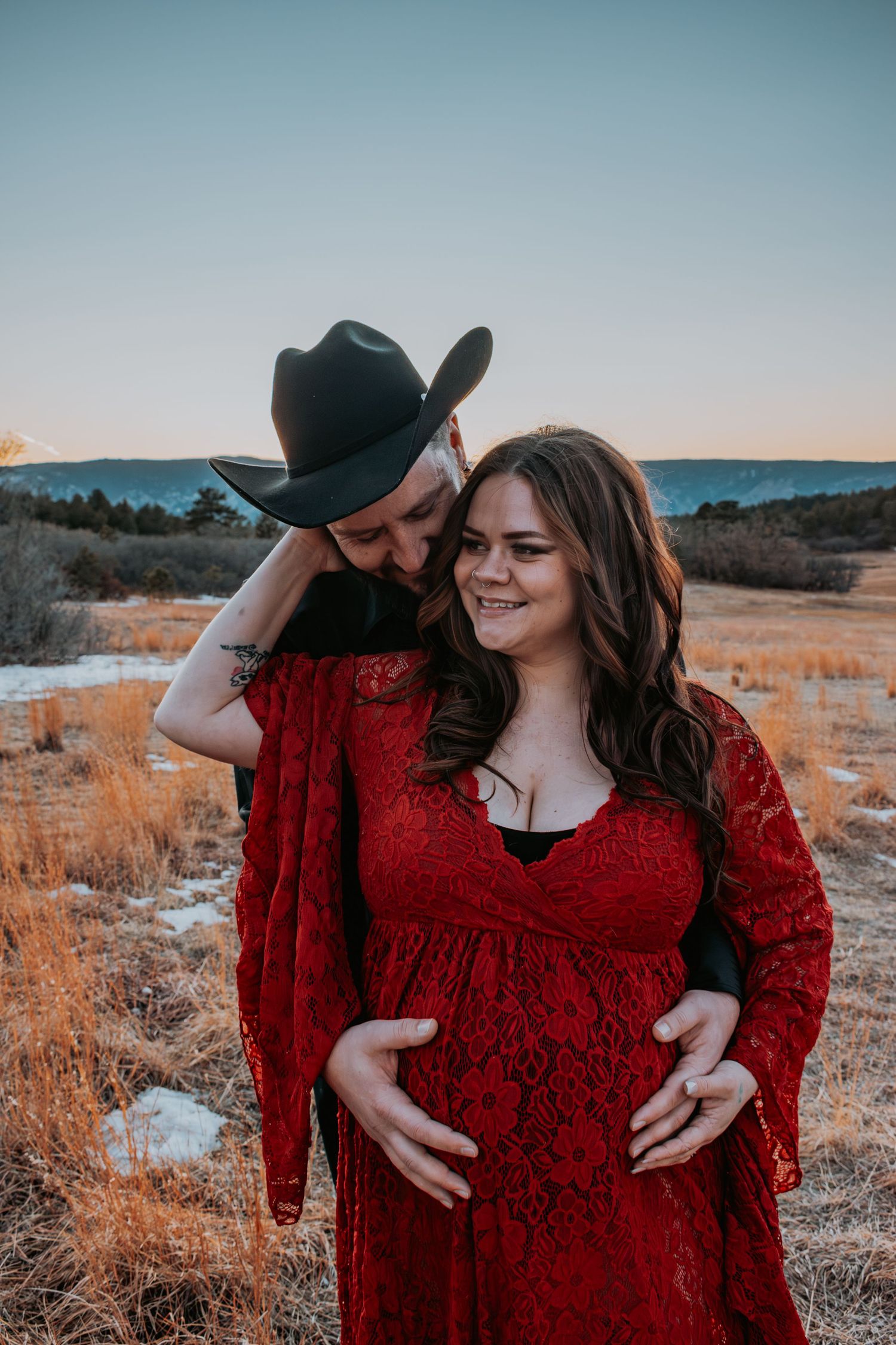 Pregnant couple in red lace maternity gown and black cowboy hat embracing in golden field at sunset.
