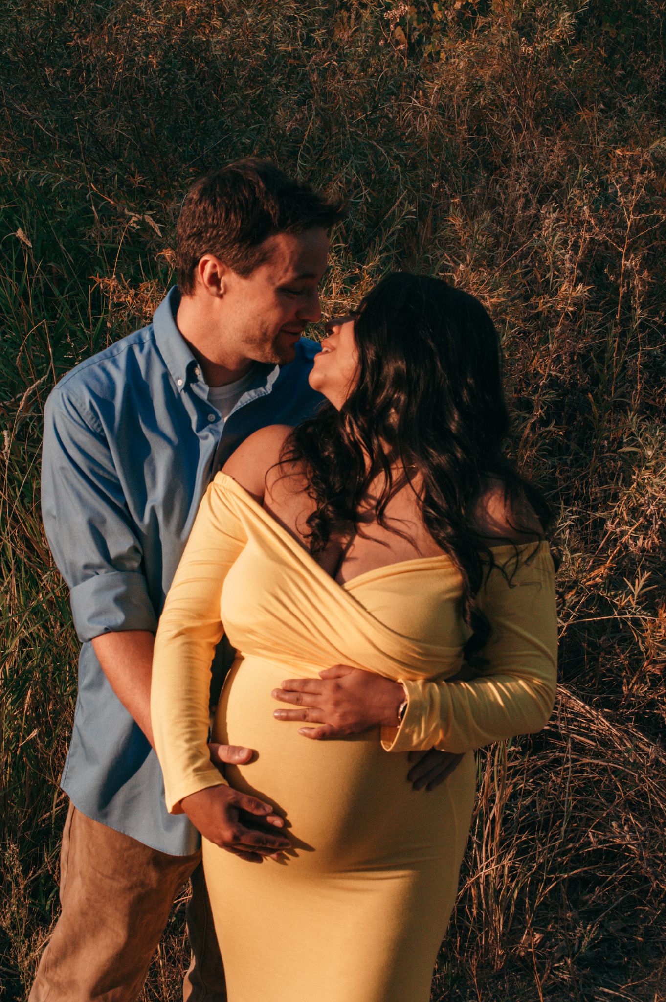 Couple sharing a tender moment outdoors, pregnant woman in yellow off-shoulder gown embraced by partner in golden field.