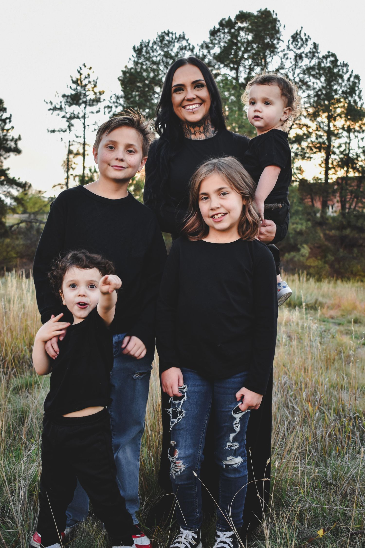 A tattooed mother smiles proudly with her four children in black outfits during an outdoor sunset photo session.