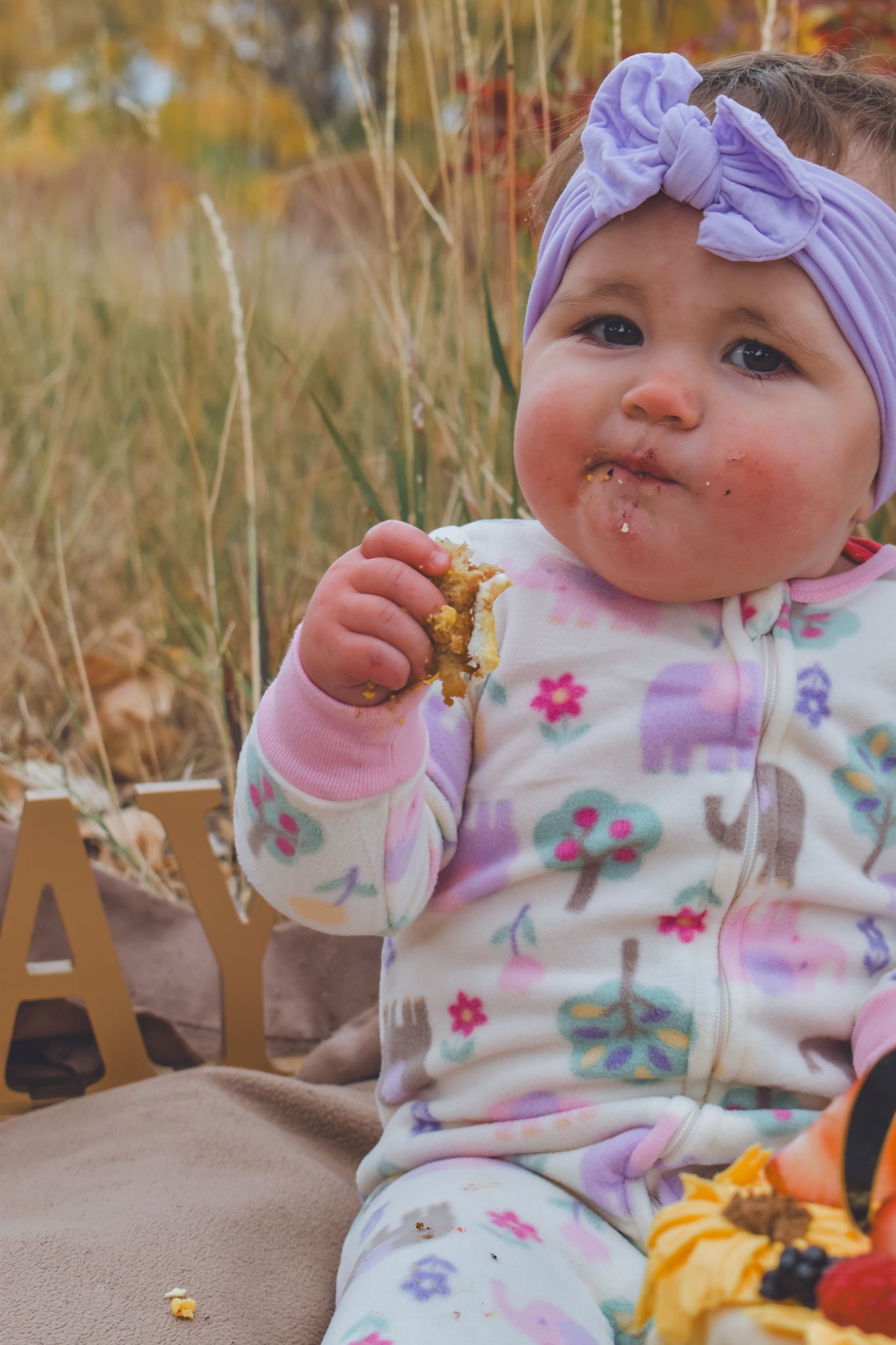 Messy baby girl with purple bow headband eating birthday cake outdoors in fall foliage setting. Family Photographer in Pflugerville, Tx