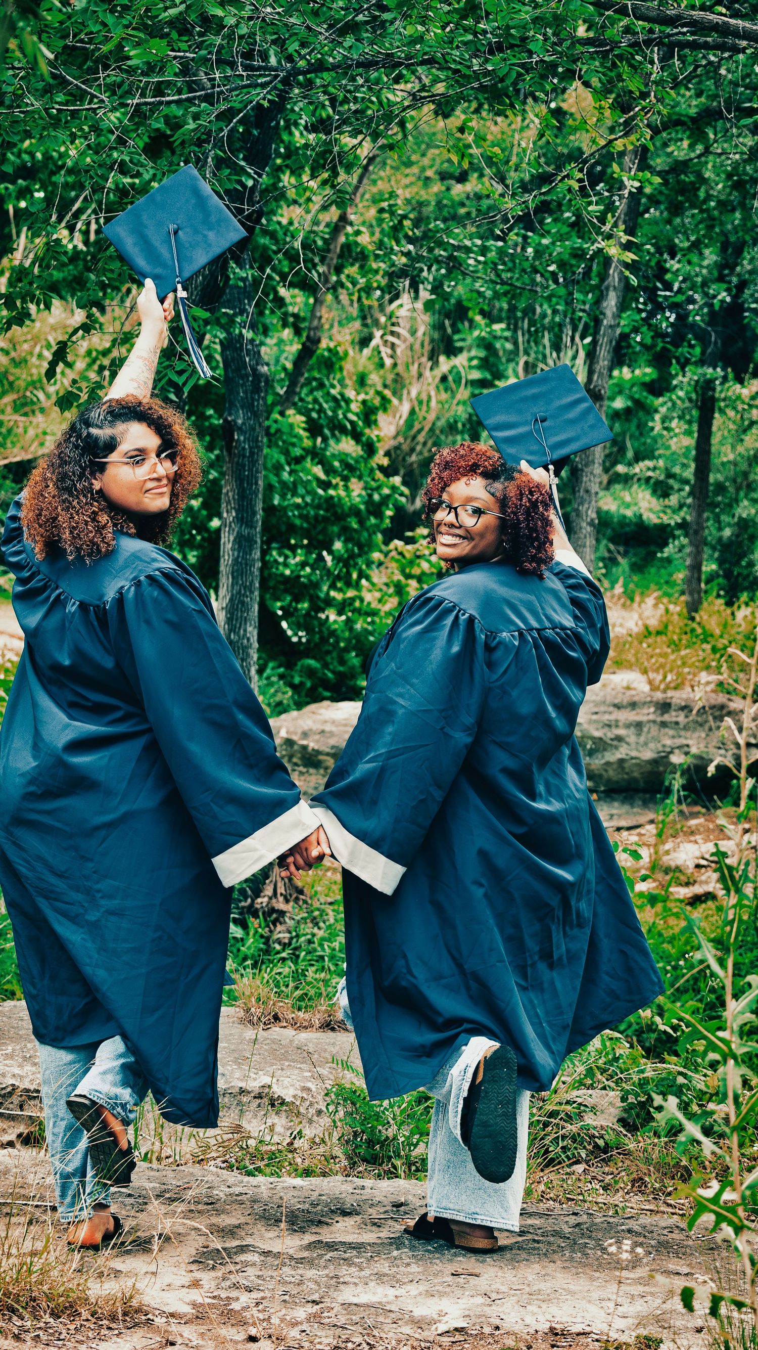 Two female graduates in blue caps and gowns celebrate outdoors among lush green trees for senior photos.