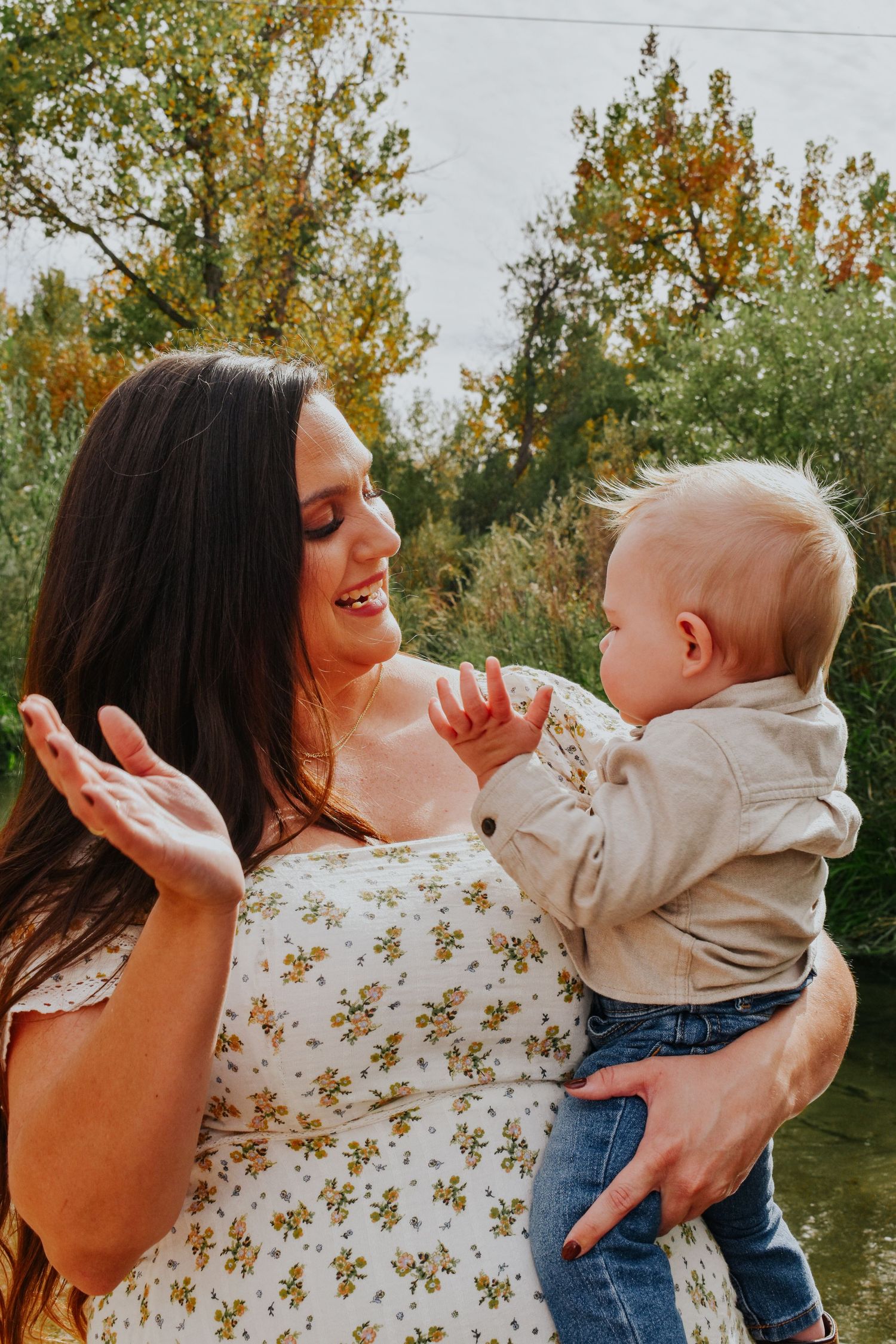 Smiling dark-haired mother in floral dress interacting playfully with her blonde baby outdoors.