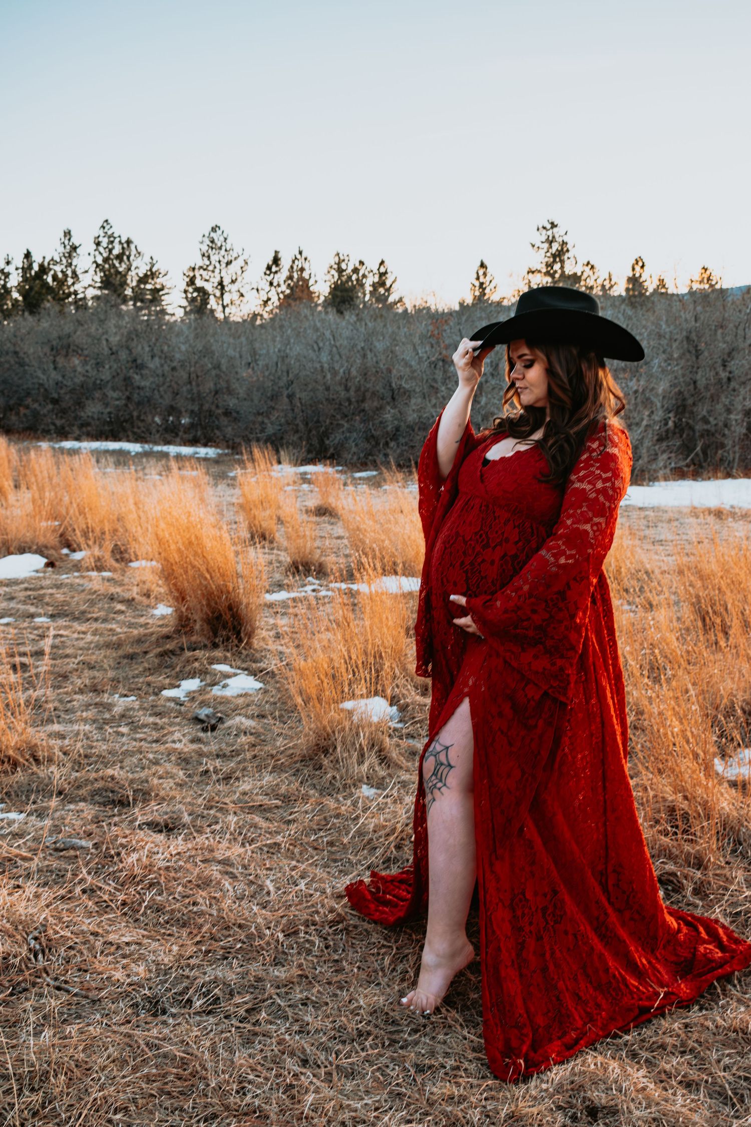 Barefoot pregnant woman in red lace gown and black cowboy hat poses in winter field.
