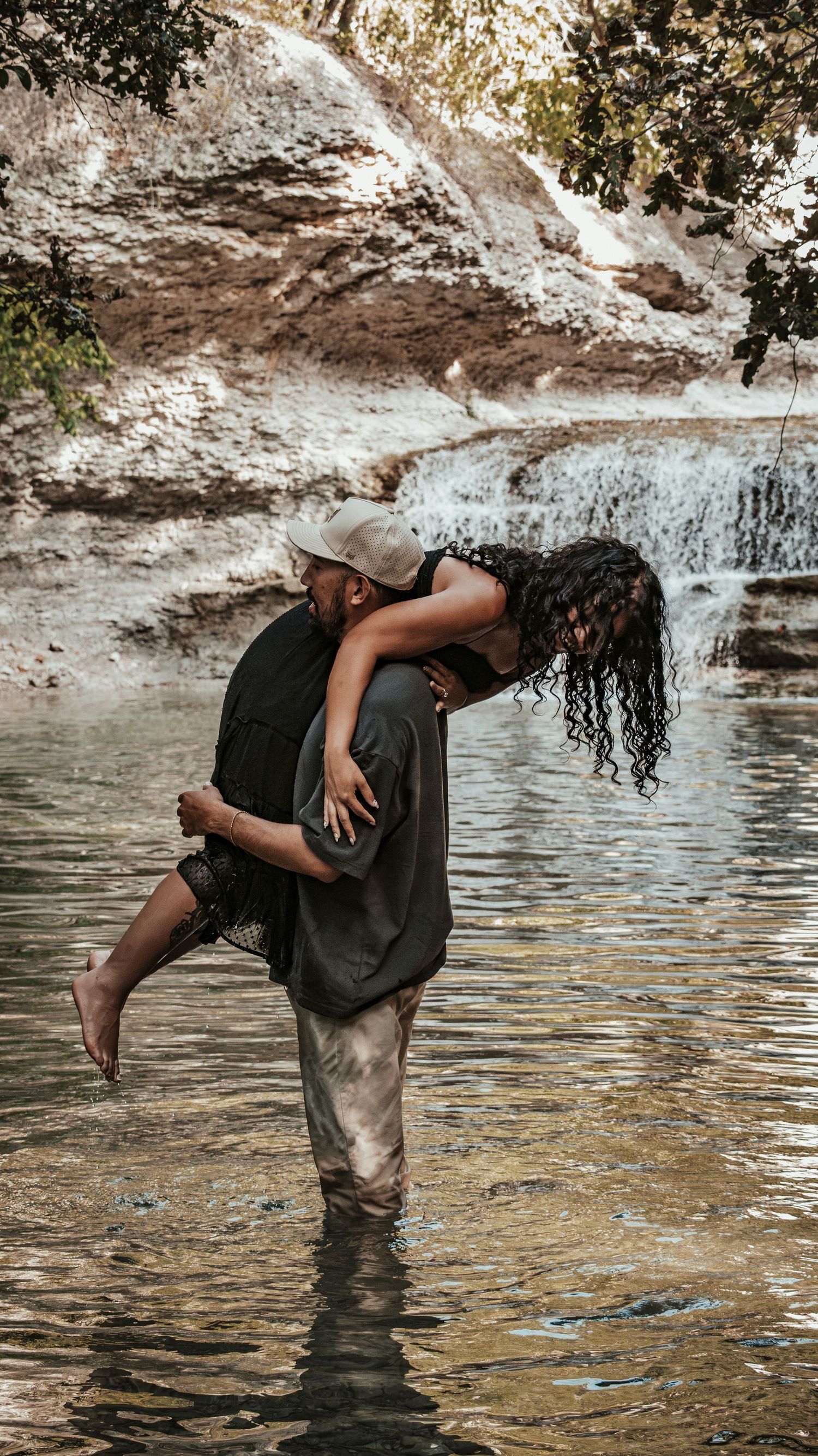 Man lifting woman in his arms while standing in a river with a waterfall backdrop.
