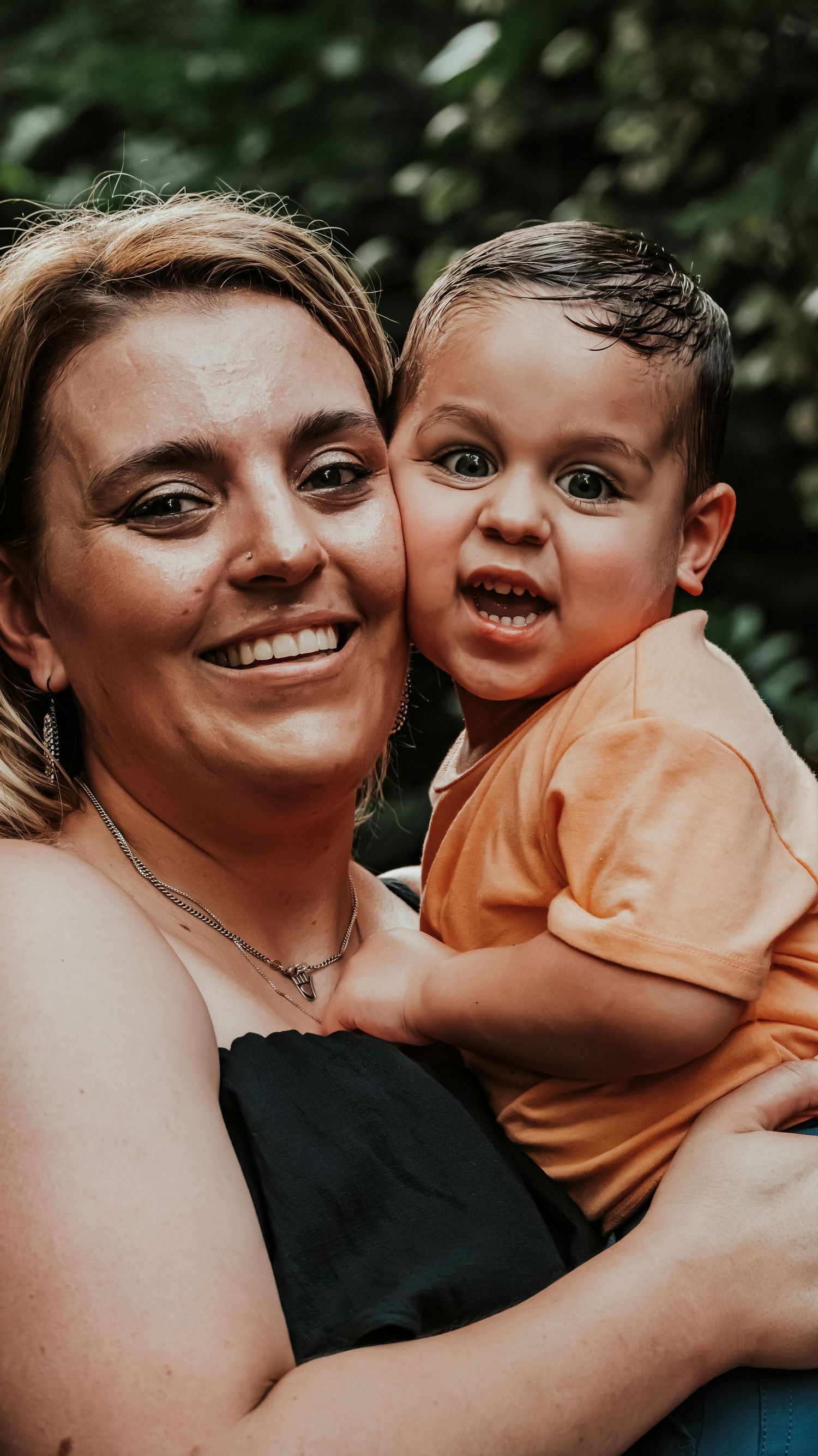 Close-up portrait of a smiling mother holding her expressive toddler son in an orange shirt outside.