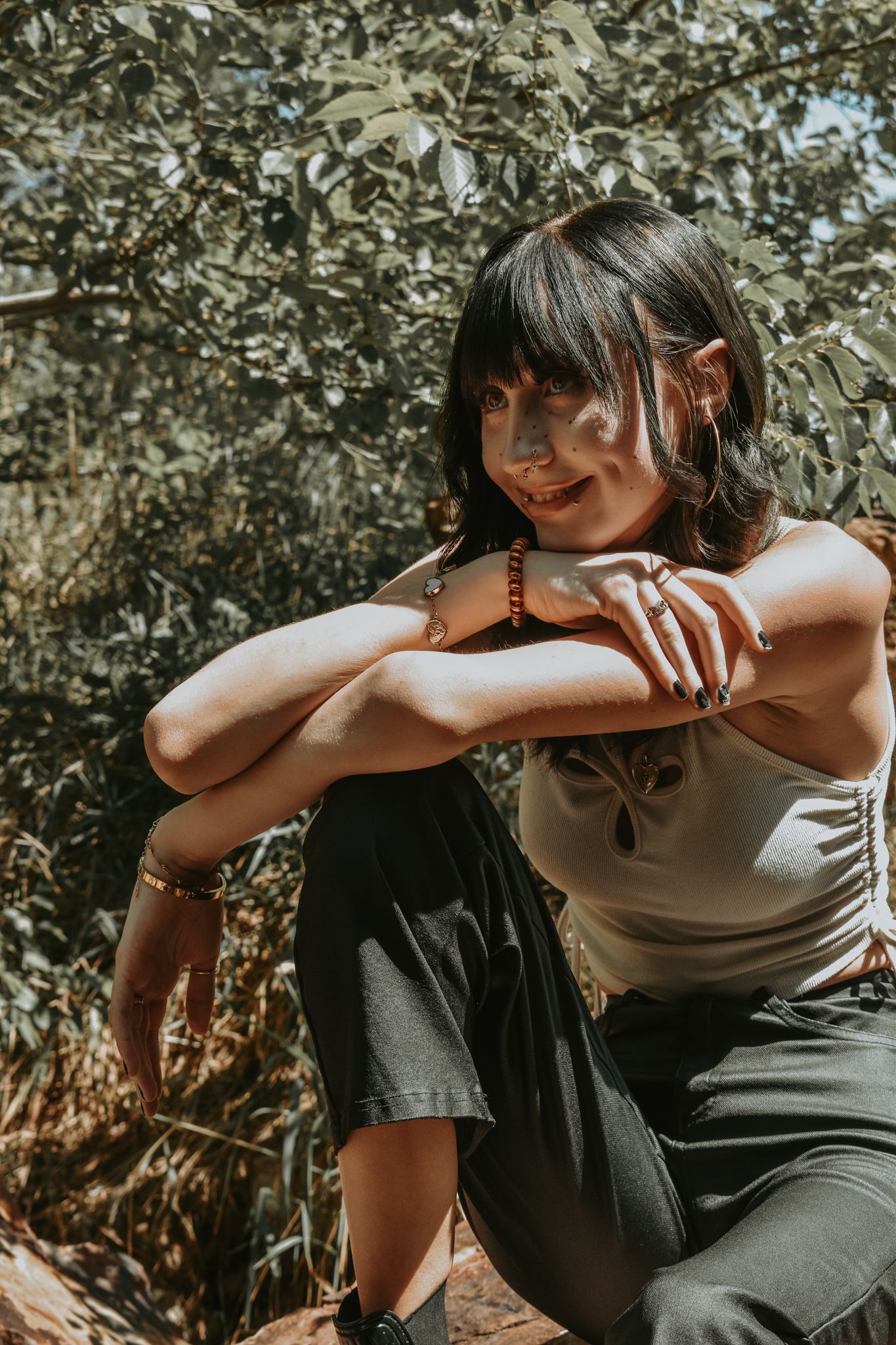 Dark-haired woman in white tank top and black pants sits outdoors in dappled sunlight among green foliage for senior photos.