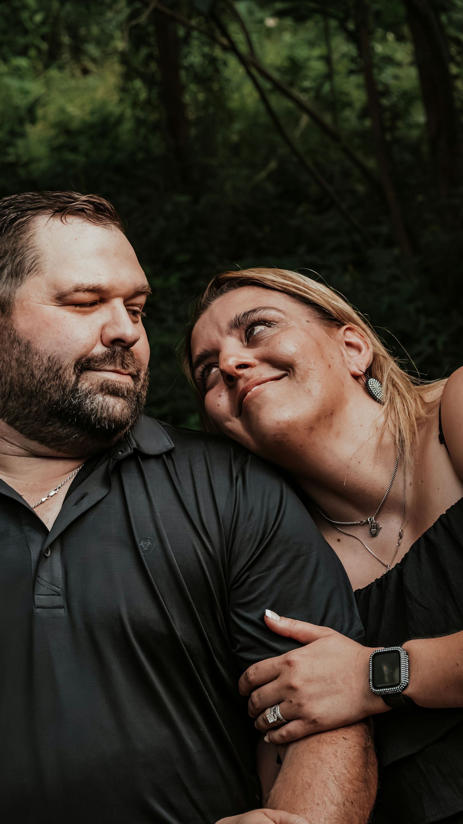 Couple sharing a tender moment outdoors, woman smiling while leaning on mans shoulder in green forest.