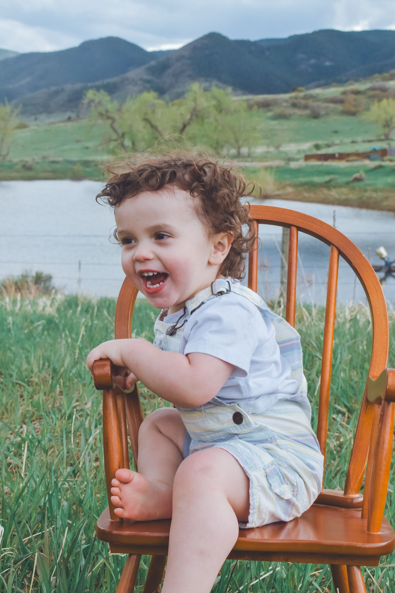 Happy laughing toddler in overalls sitting in a wooden chair with a lake and mountains behind. Family Photographer in Pflugerville, Tx