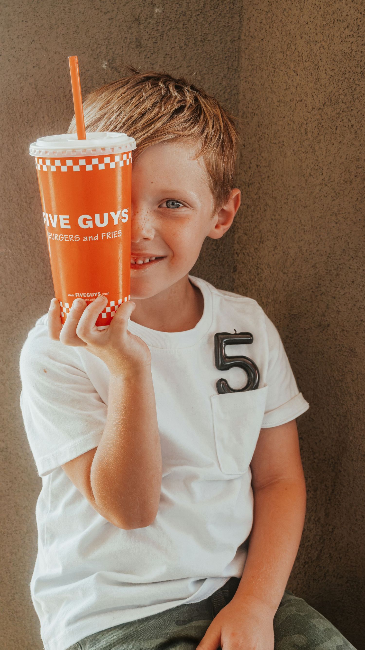 Smiling boy holding an orange Five Guys cup with a straw, wearing a white shirt with a number 5. Family Photographer in Pflugerville, Tx