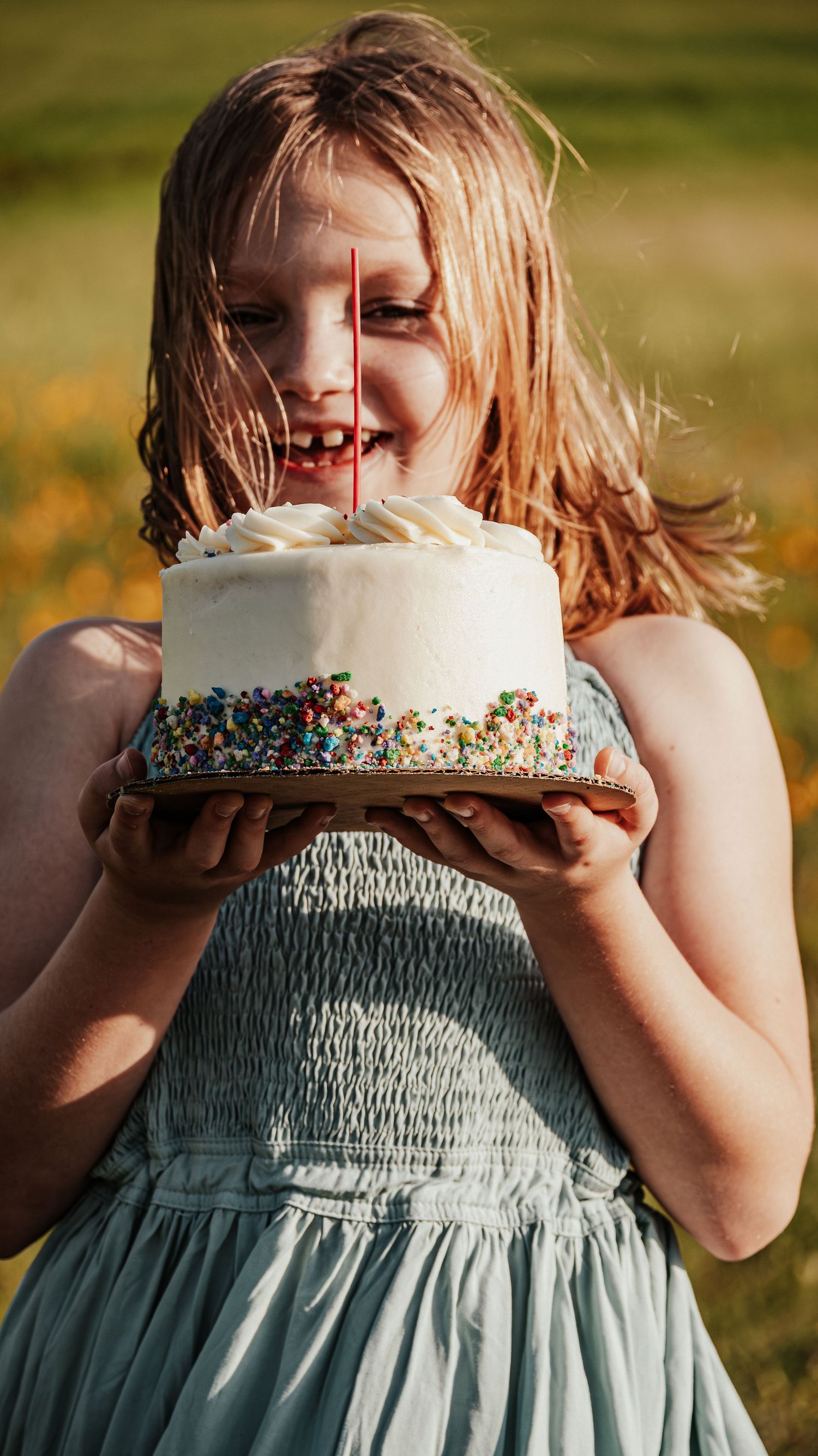 Smiling girl holding white birthday cake with sprinkles and candles outdoors.
