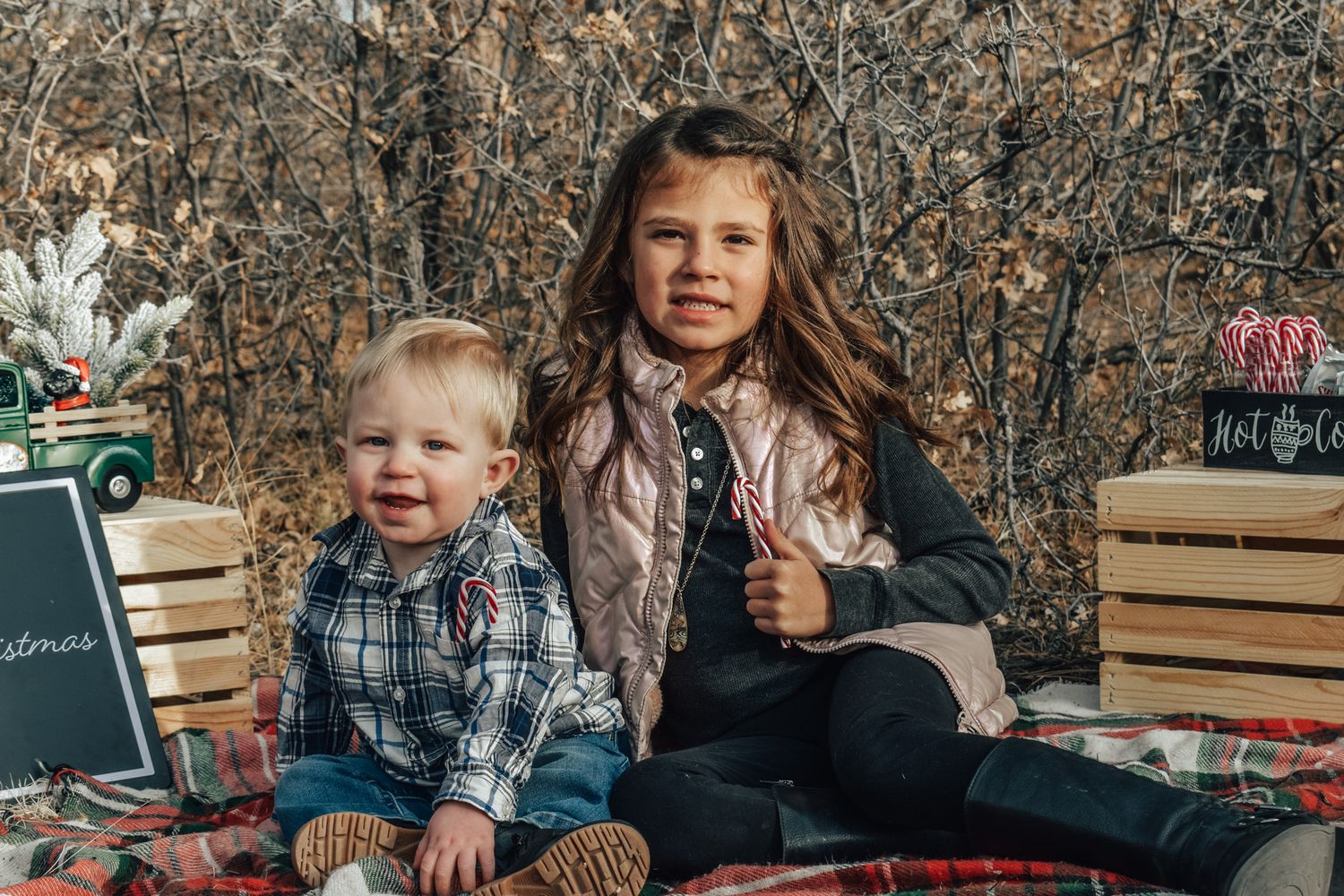A young girl and toddler boy pose together on a red plaid blanket with festive Christmas decorations outdoors.