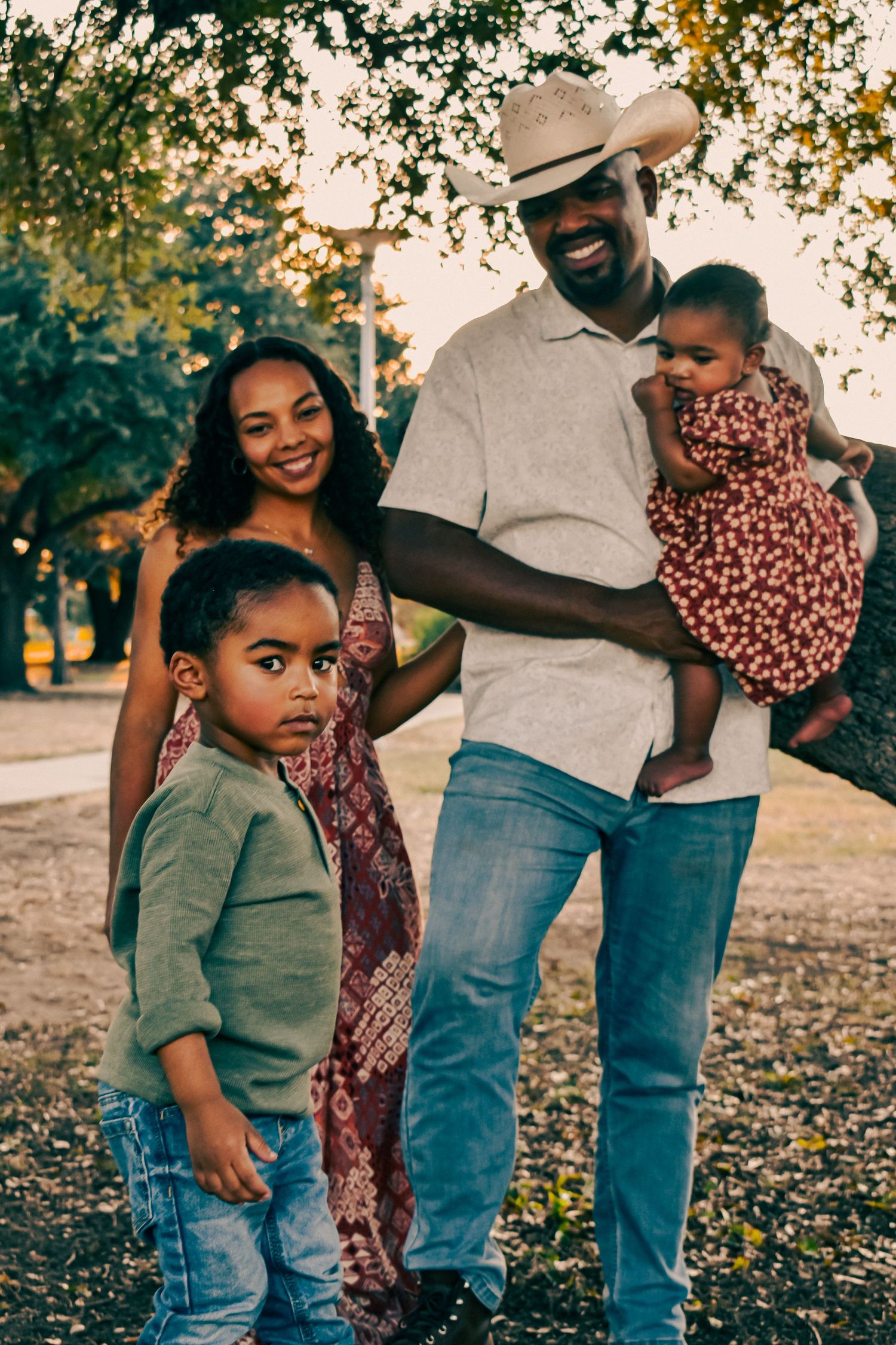 Happy African American family of four poses under a tree at sunset, dad in cowboy hat holding baby girl with son and mom.