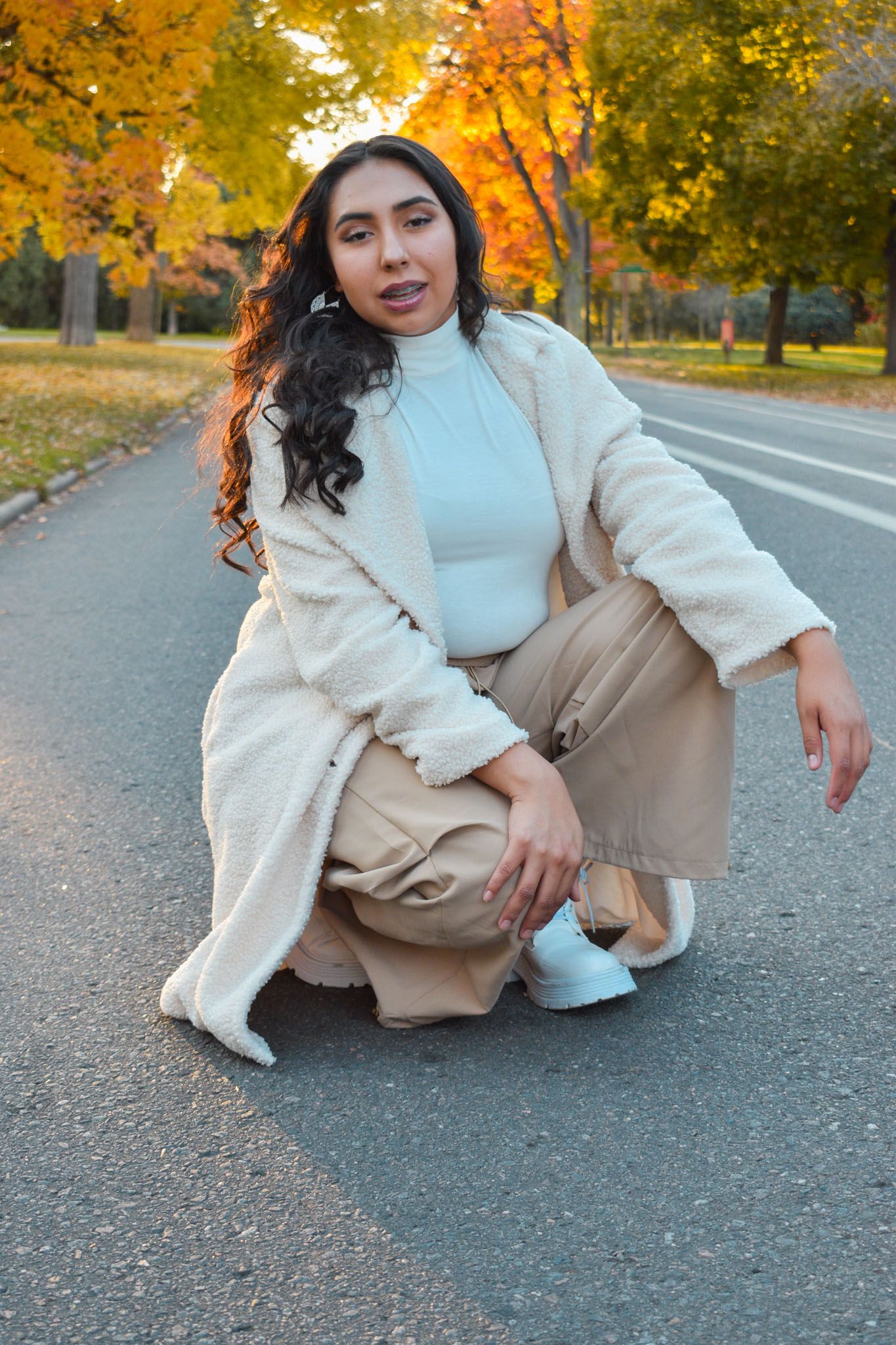 Young woman in beige coat and tan pants crouching on road surrounded by colorful autumn trees for senior photos.