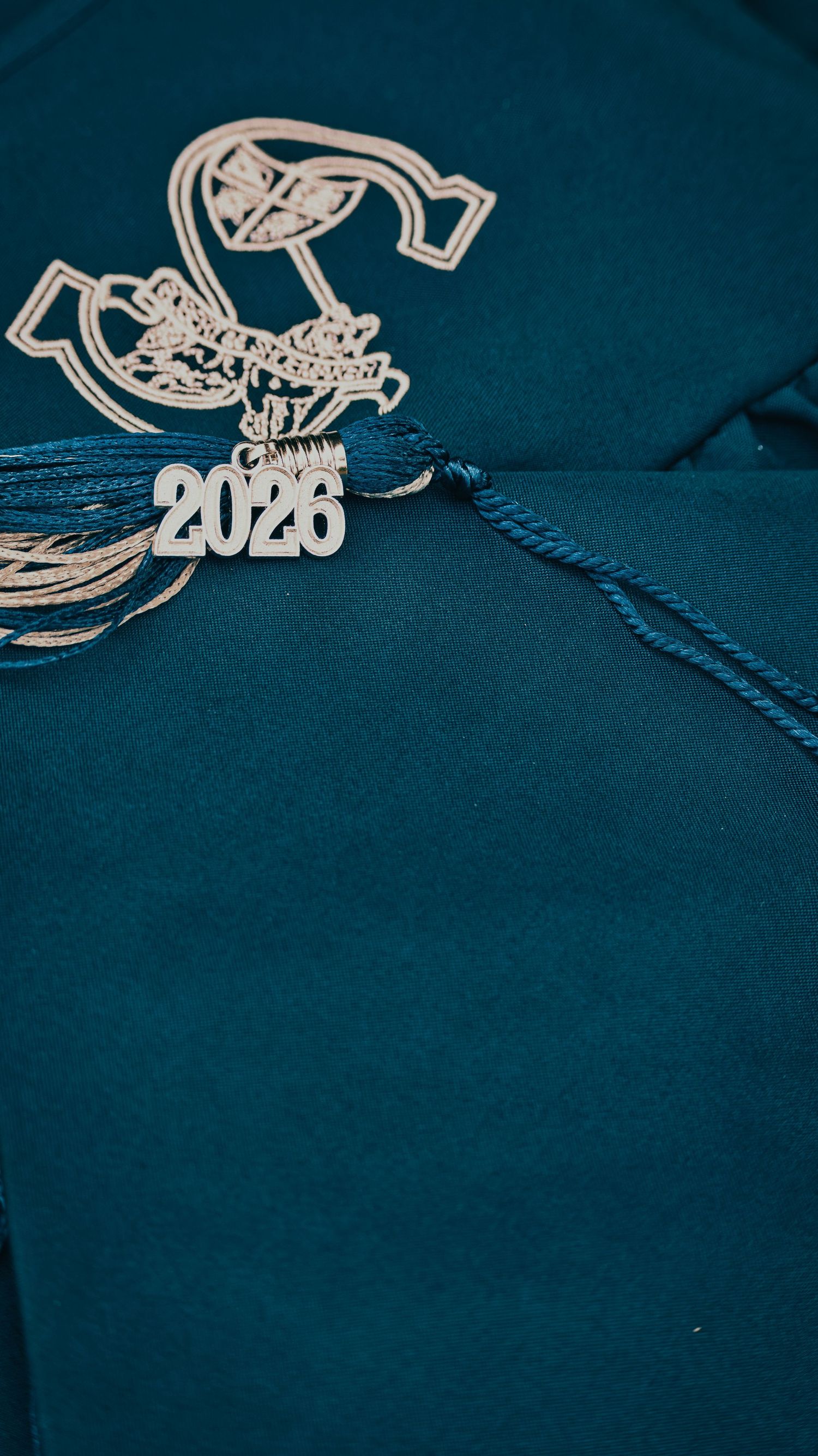 Teal graduation cap with gold emblem and blue tassel featuring a 2026 charm for senior photos.