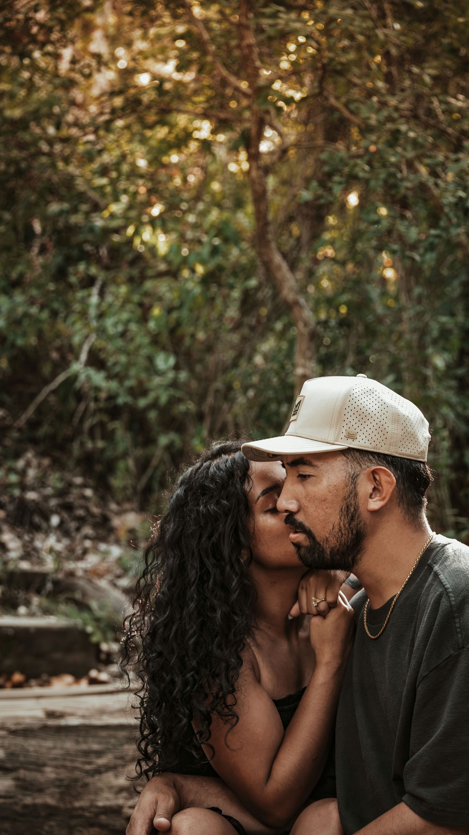Romantic couple sharing a kiss outdoors surrounded by lush green trees in natural light.
