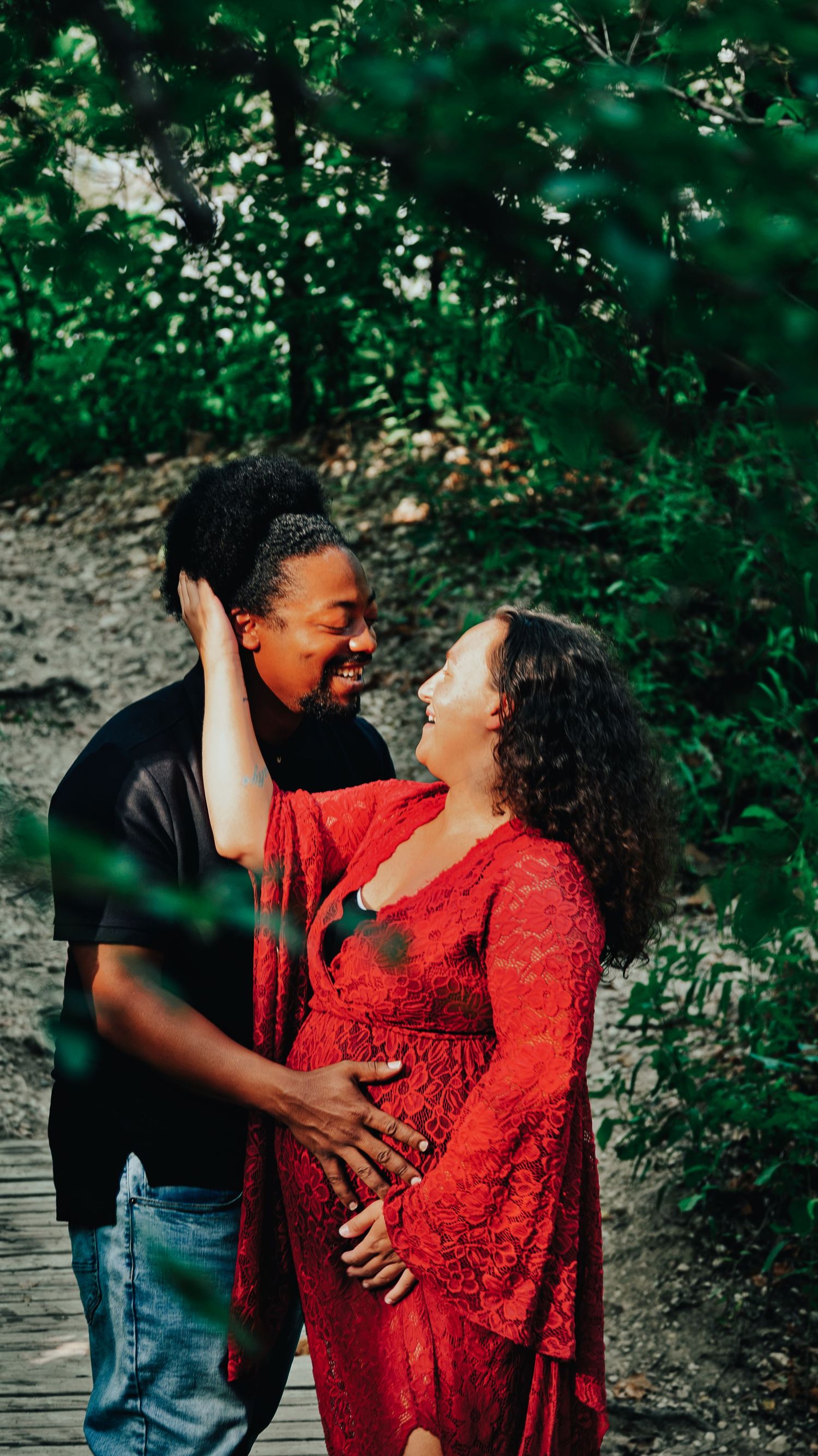 Smiling couple embracing outdoors, man holding pregnant womans belly in a red lace maternity gown.