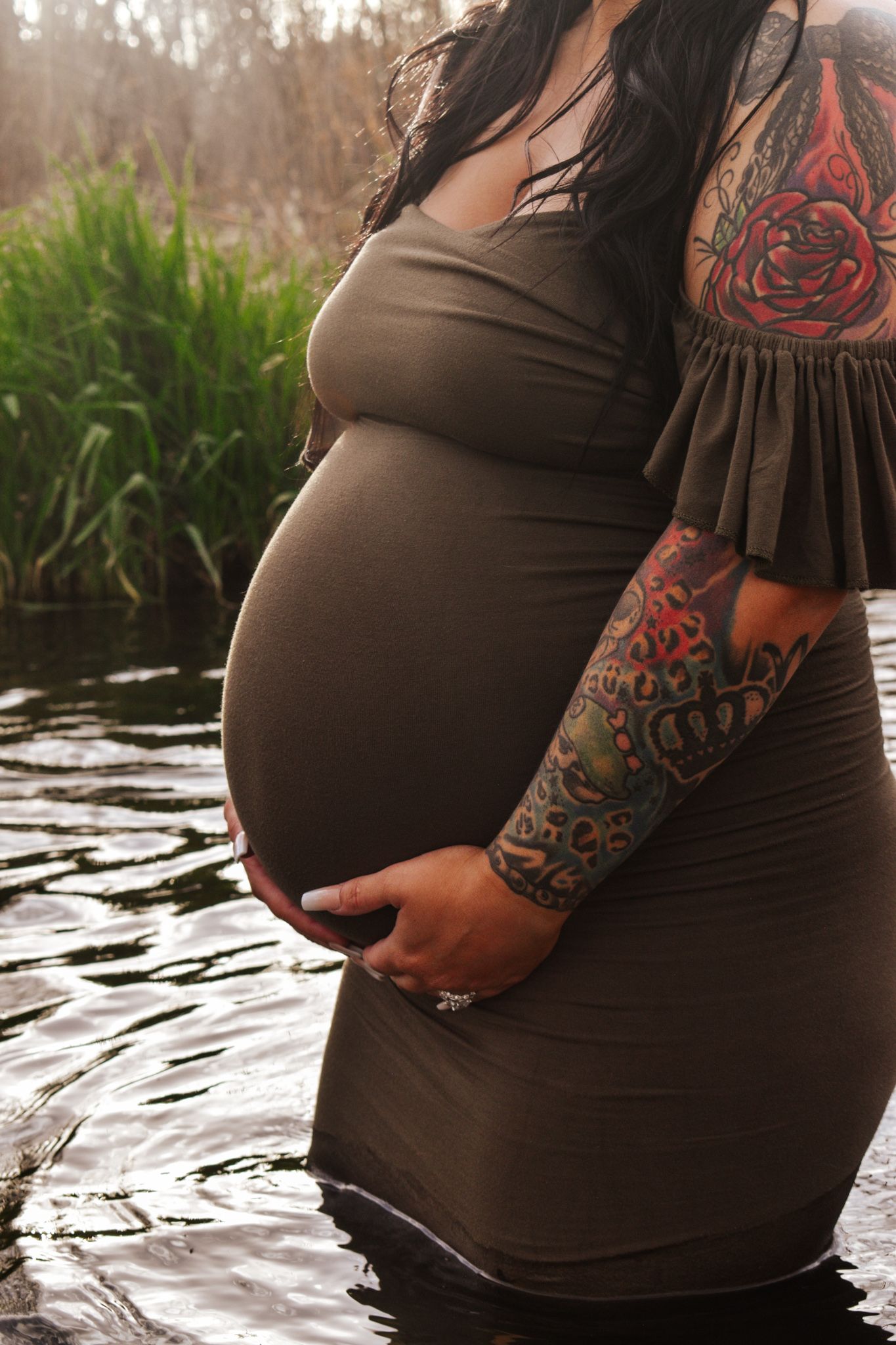 Close-up of pregnant woman in gray dress standing in river water, displaying colorful arm tattoos.