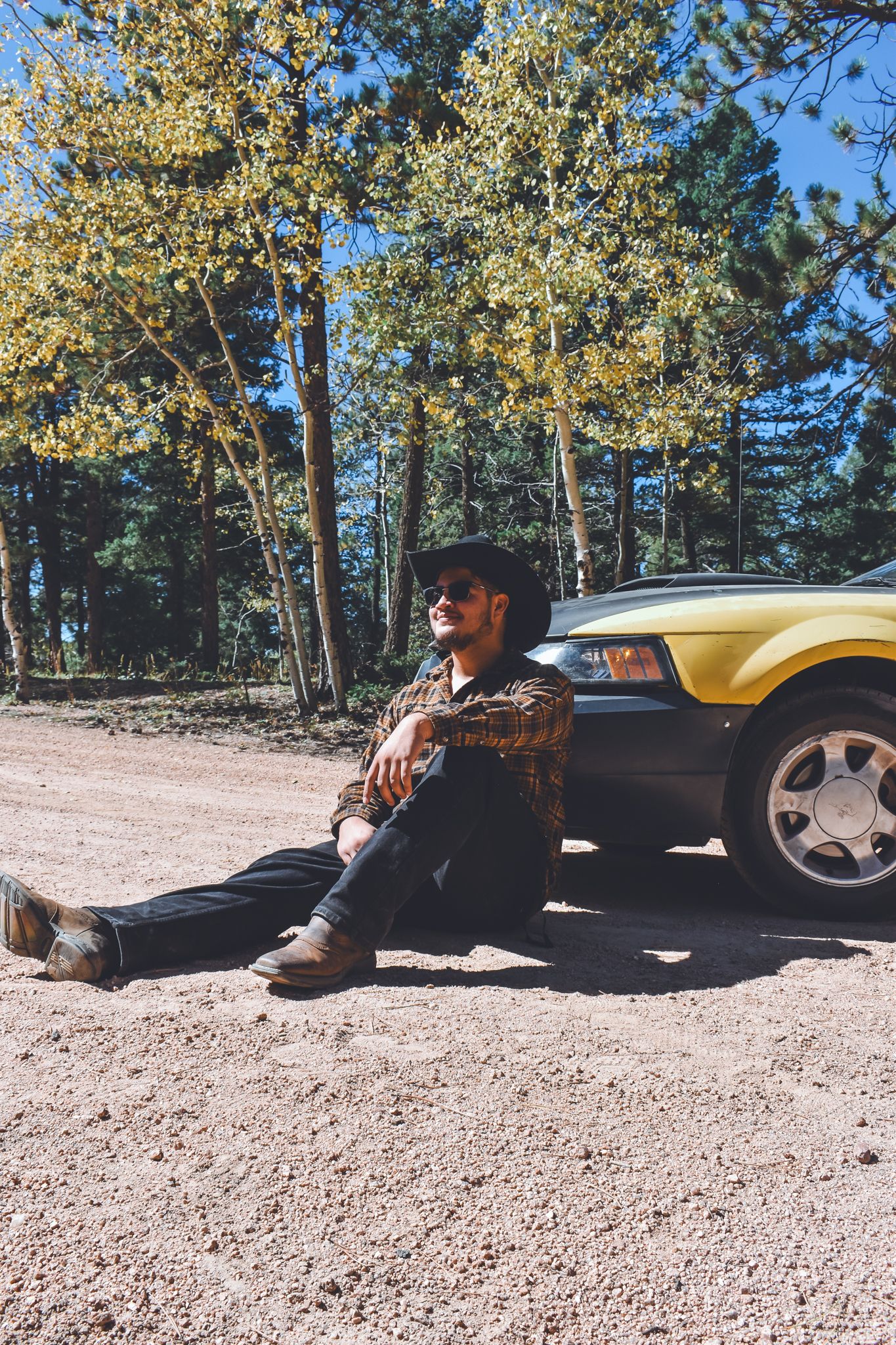 Man in cowboy hat sitting against a yellow car on a dirt road in a pine forest for senior photos.