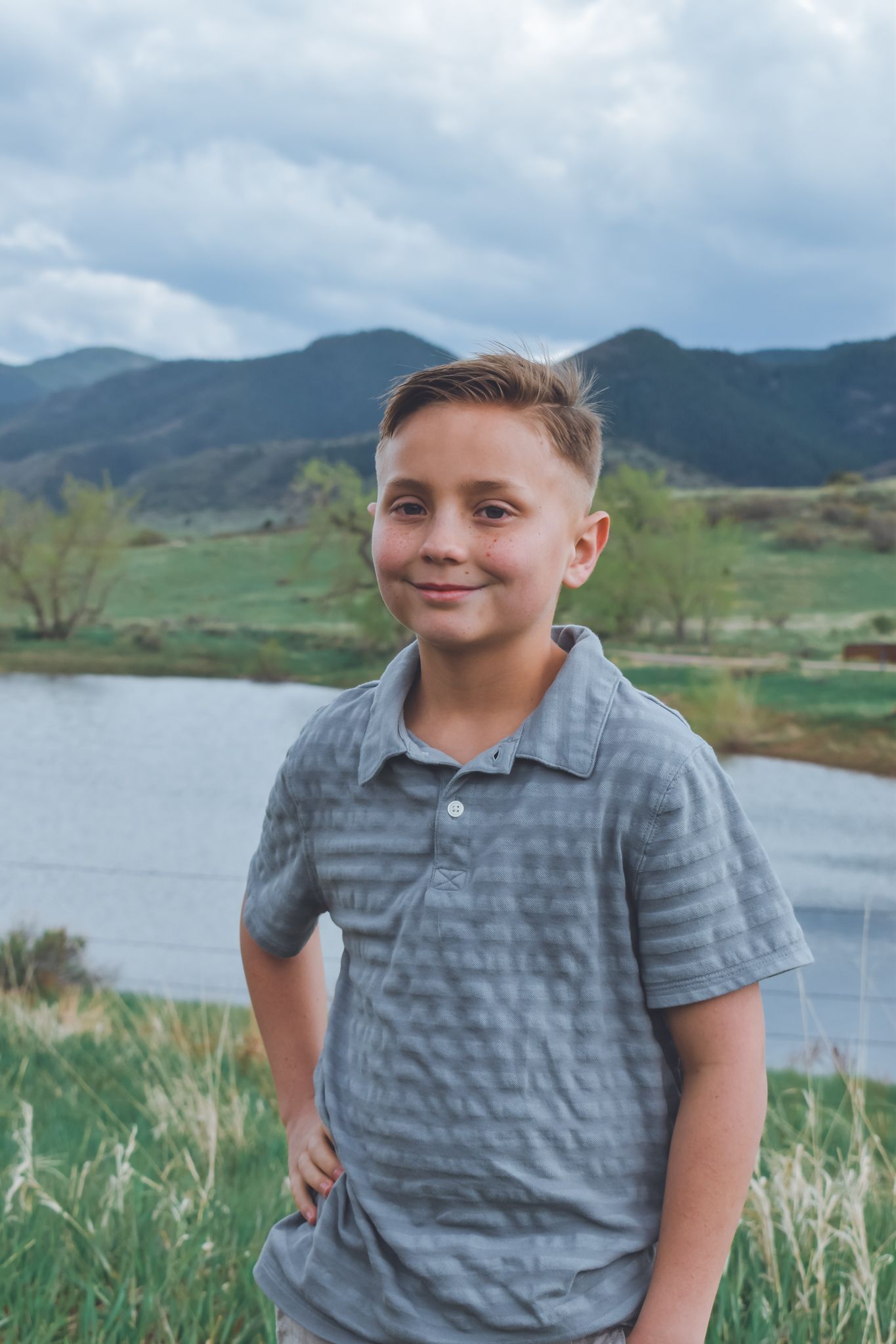 Smiling young boy in gray polo shirt standing near mountain lake on cloudy day.