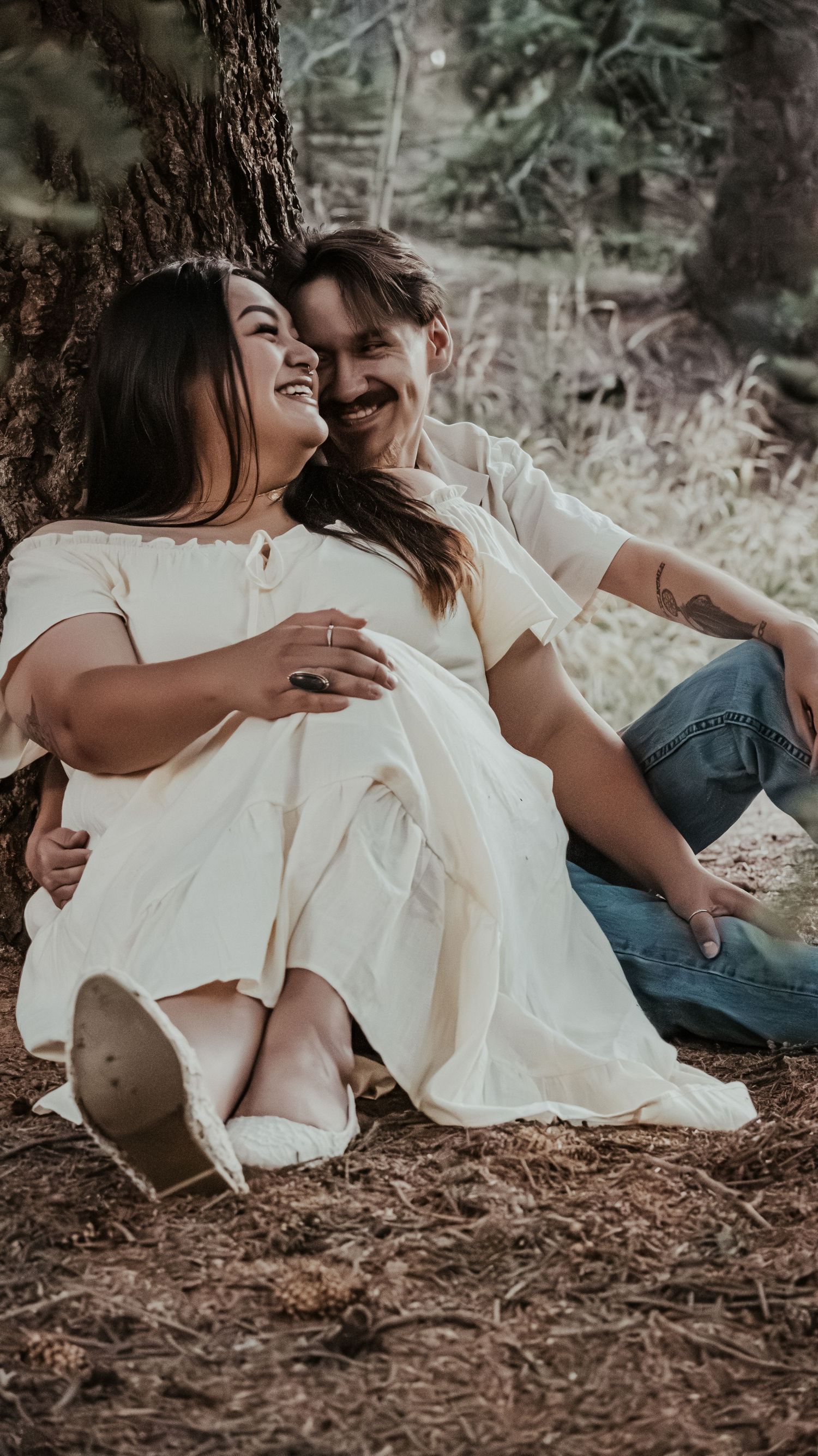 A happy couple sits together against a tree, the woman in a cream dress resting on her partners lap.