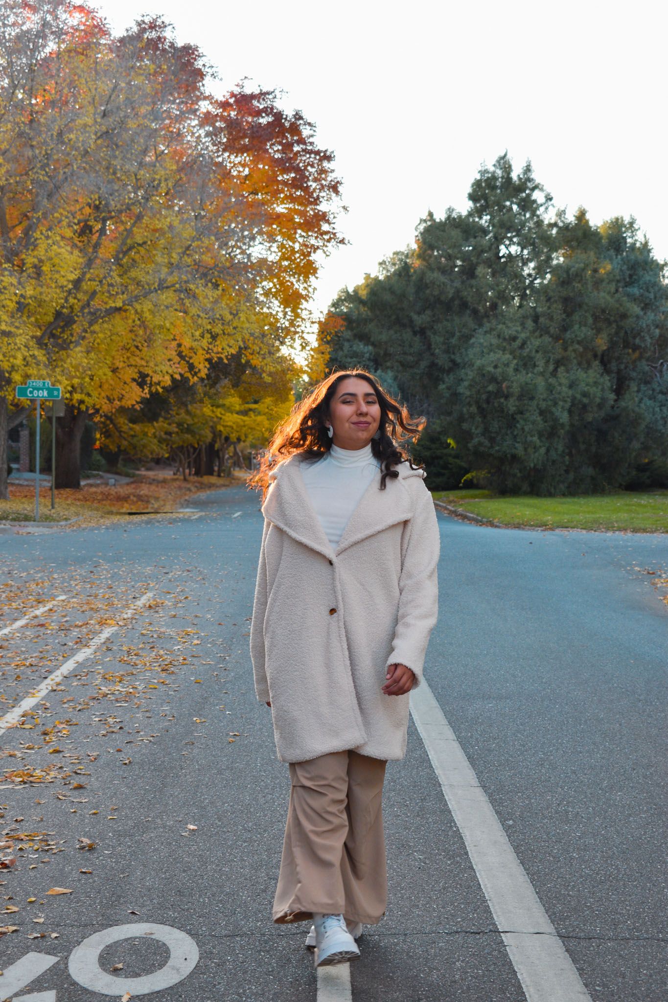 Woman in cream coat walking down road lined with vibrant orange and yellow autumn trees for senior photos.