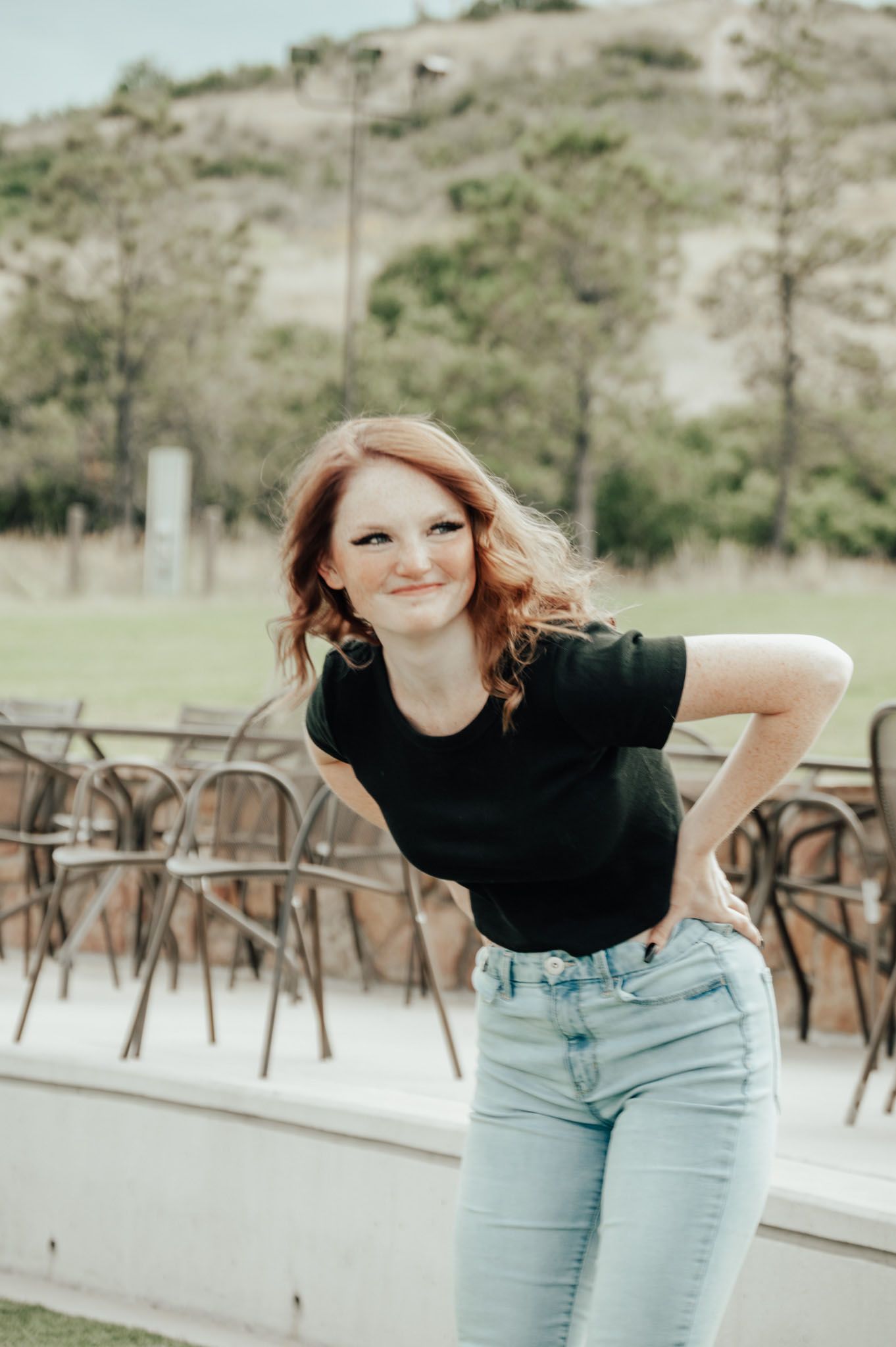 Smiling redhead woman in black tee and light jeans posing outdoors with hands on hips near patio chairs for senior photos.