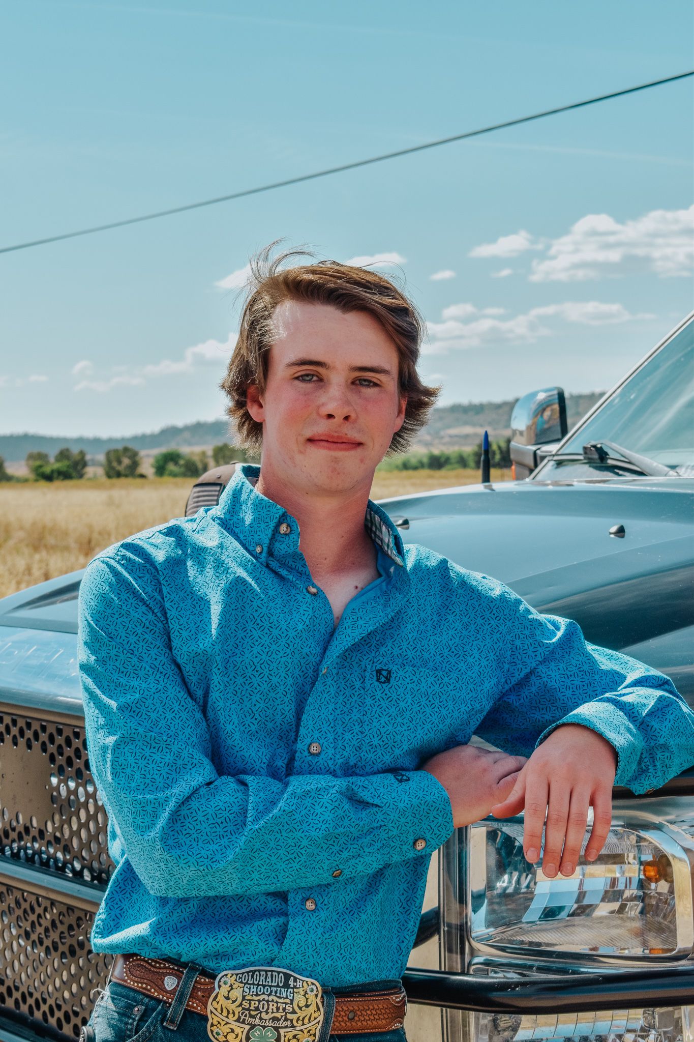 Young man in blue western shirt leans against truck in rural field under blue sky for senior photos.