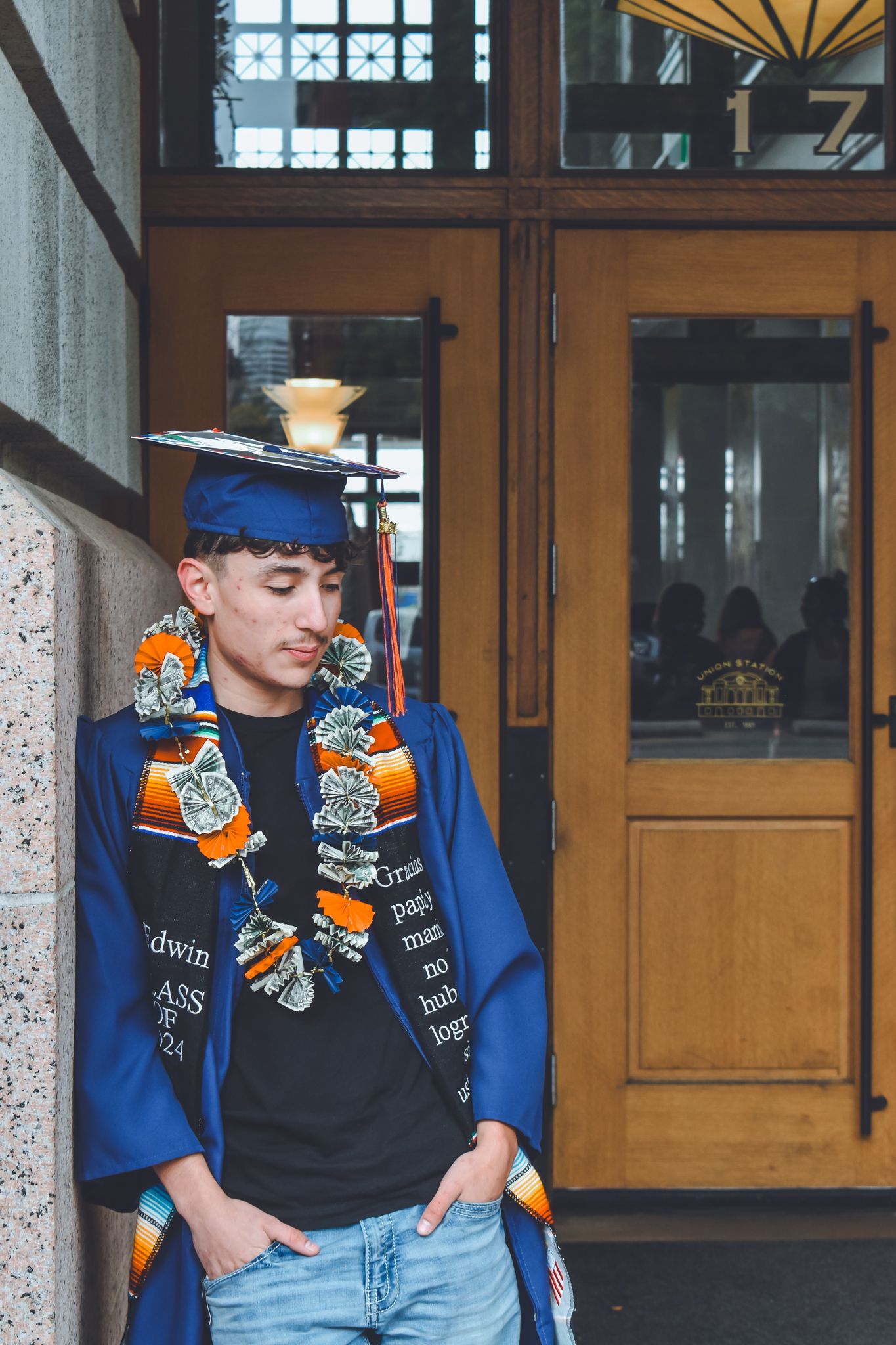 Graduate in blue cap and gown leans against wall outside historic building entrance for senior photos.