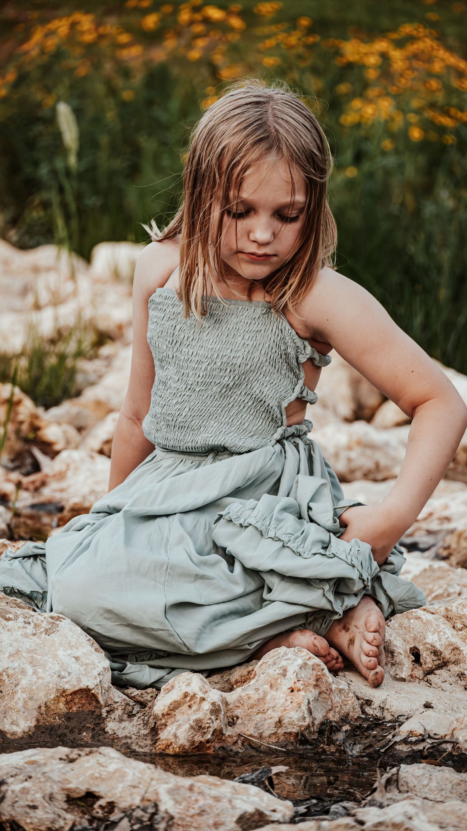 Young girl in sage green smocked top and maxi skirt sitting on rocks near water.