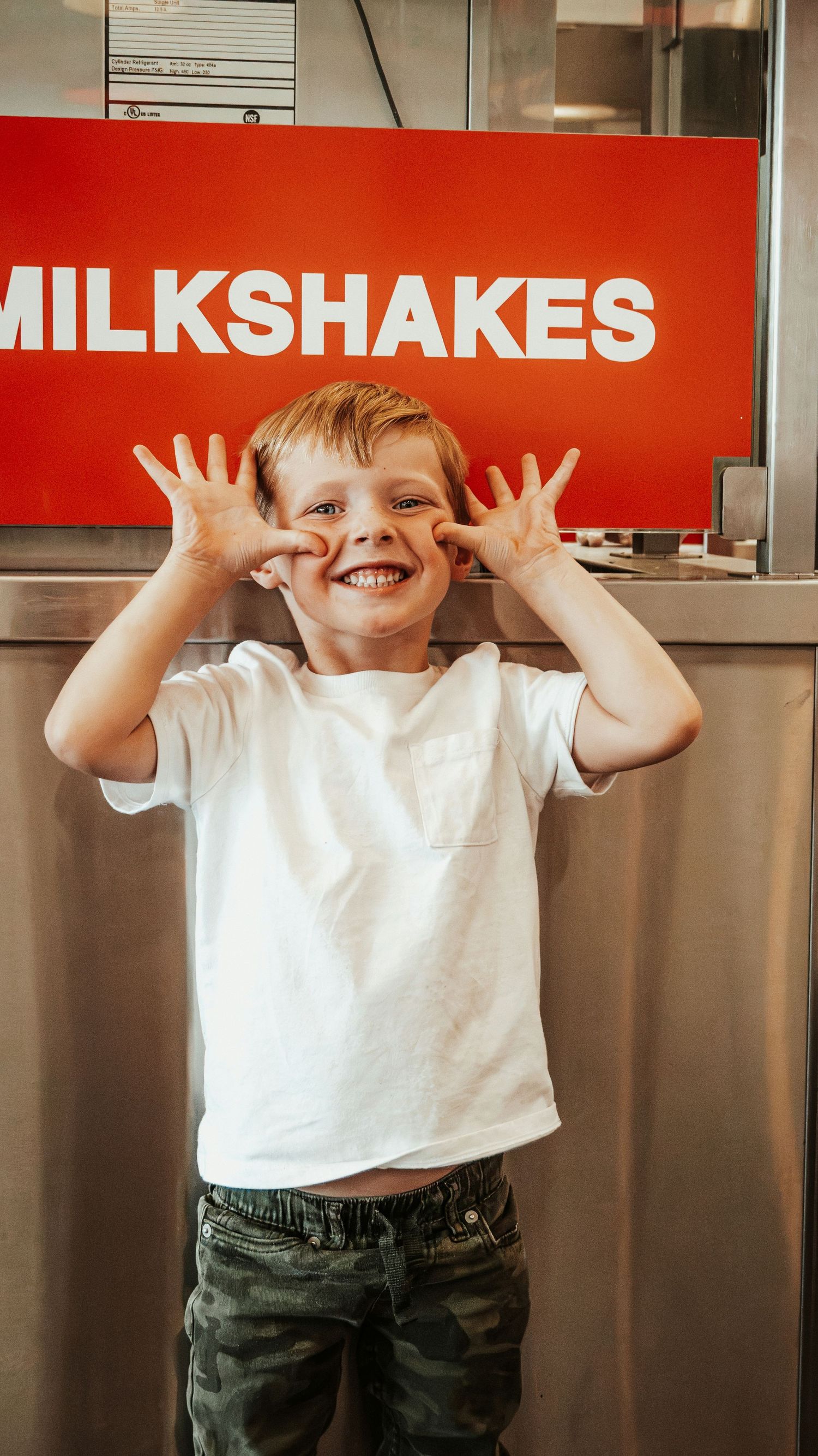 Excited boy making funny faces in front of Five Guys milkshakes sign.
