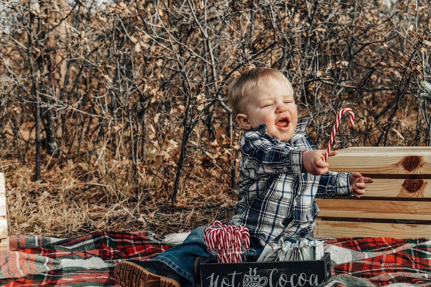 A laughing toddler boy in a plaid shirt holds a candy cane while sitting on a festive holiday blanket.