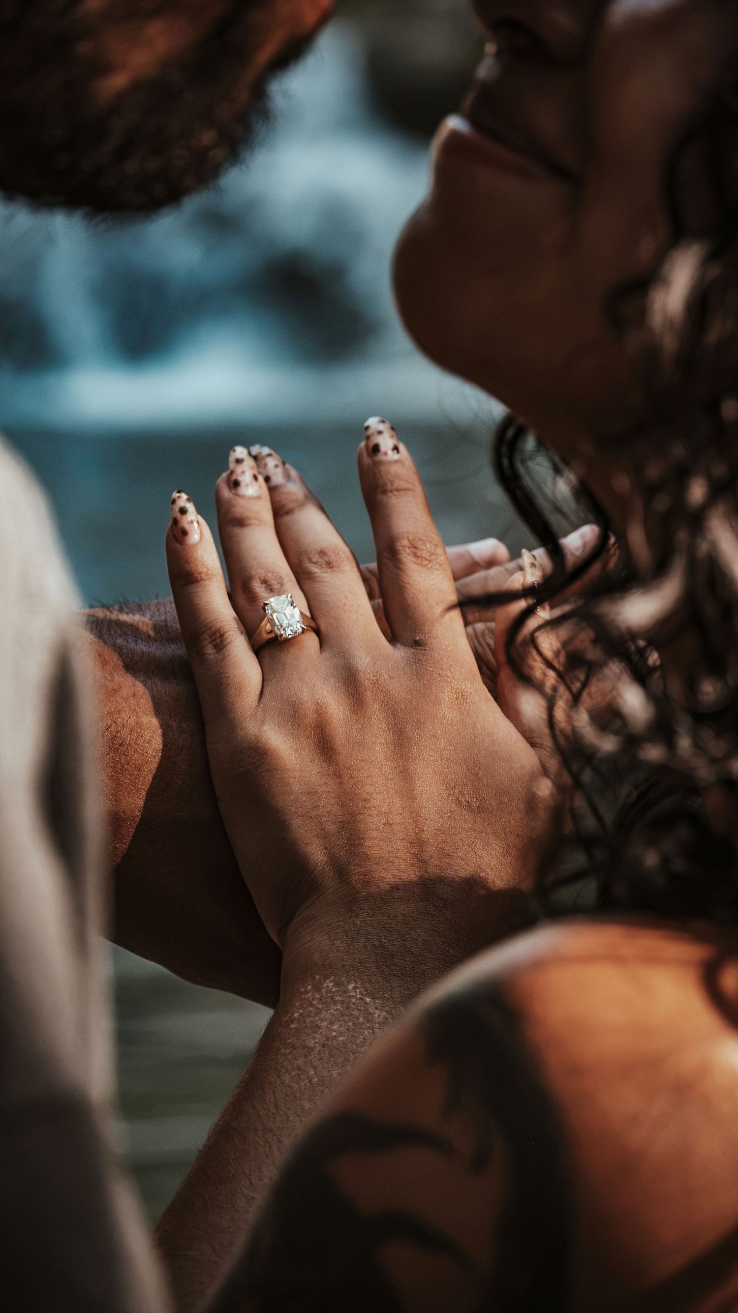 Close-up of womans hand displaying a stunning diamond engagement ring while couple shares a romantic moment near waterfall.