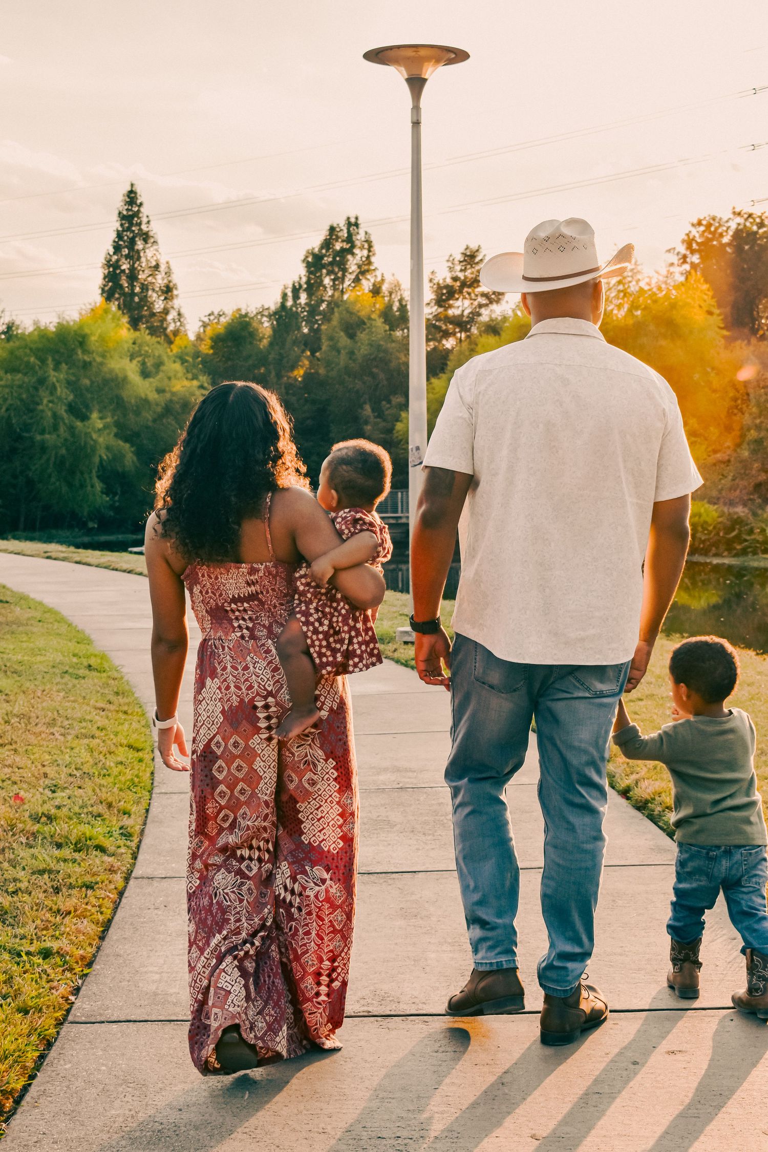 African American family strolling on a park pathway at golden hour, parents with two young children in warm sunset light.