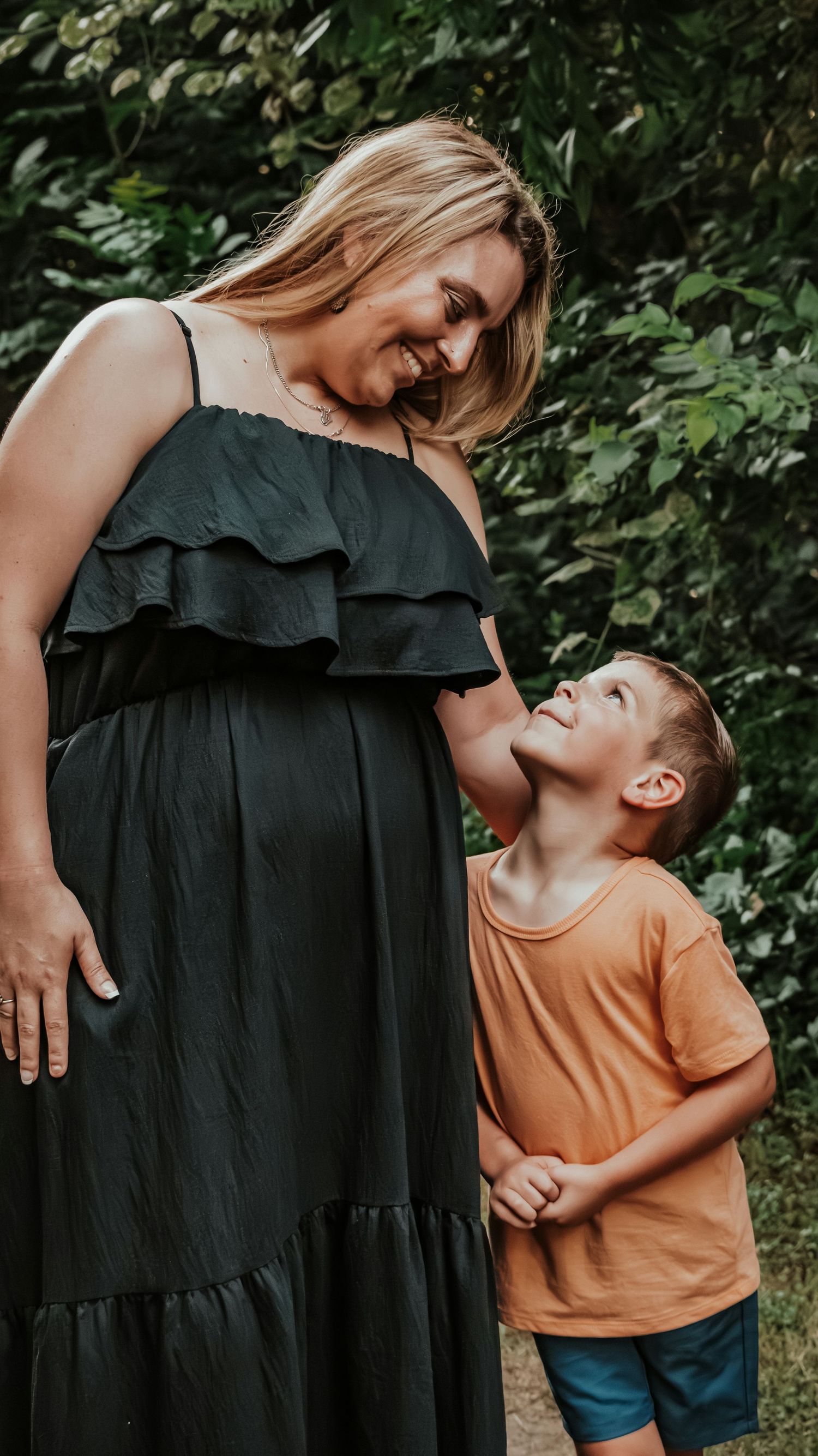 Pregnant mother in black dress sharing a joyful moment with her smiling son in an orange shirt outdoors.