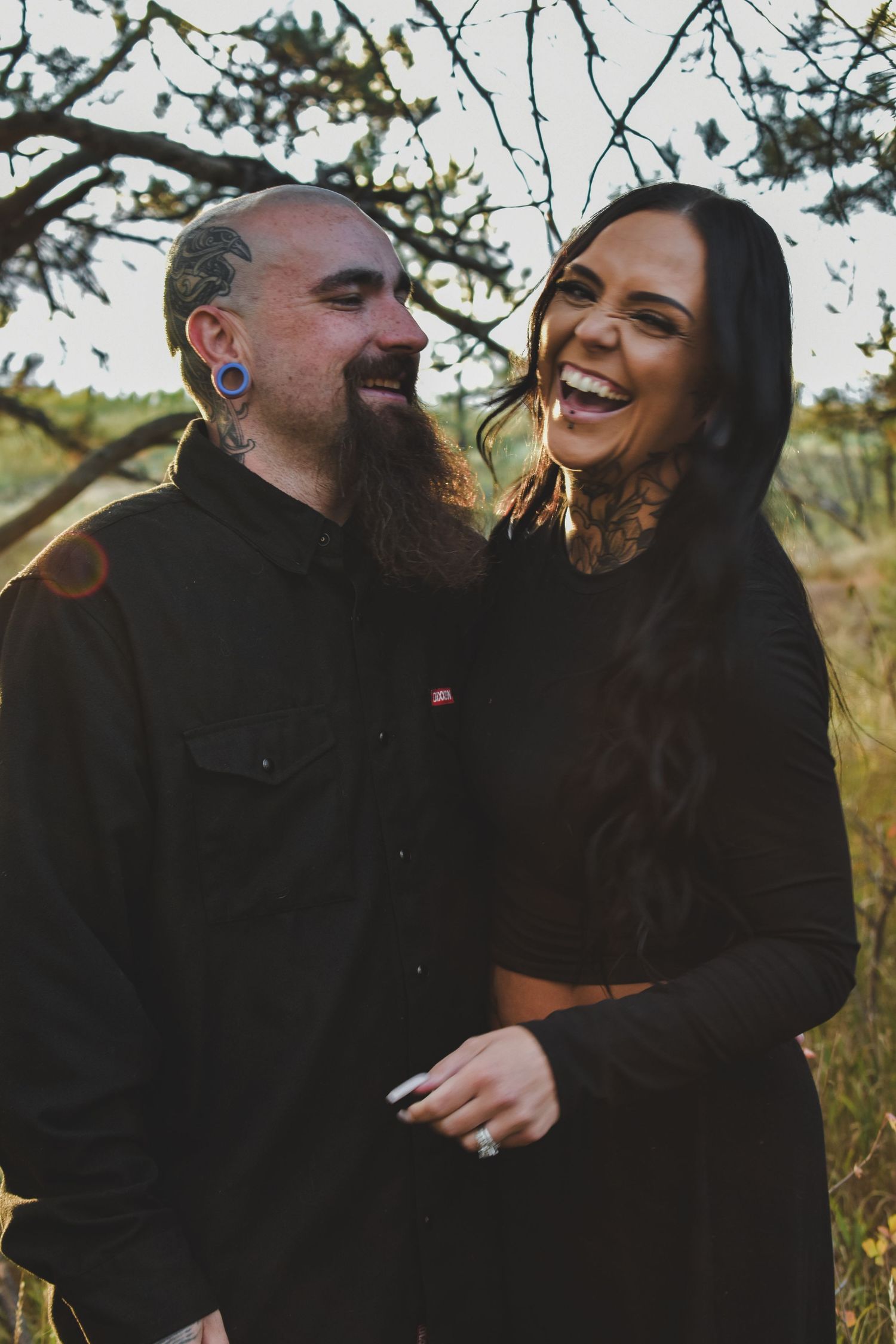 A laughing couple embraces outdoors near bare tree branches in a warm, candid engagement photo. Family Photographer in Pflugerville, Tx