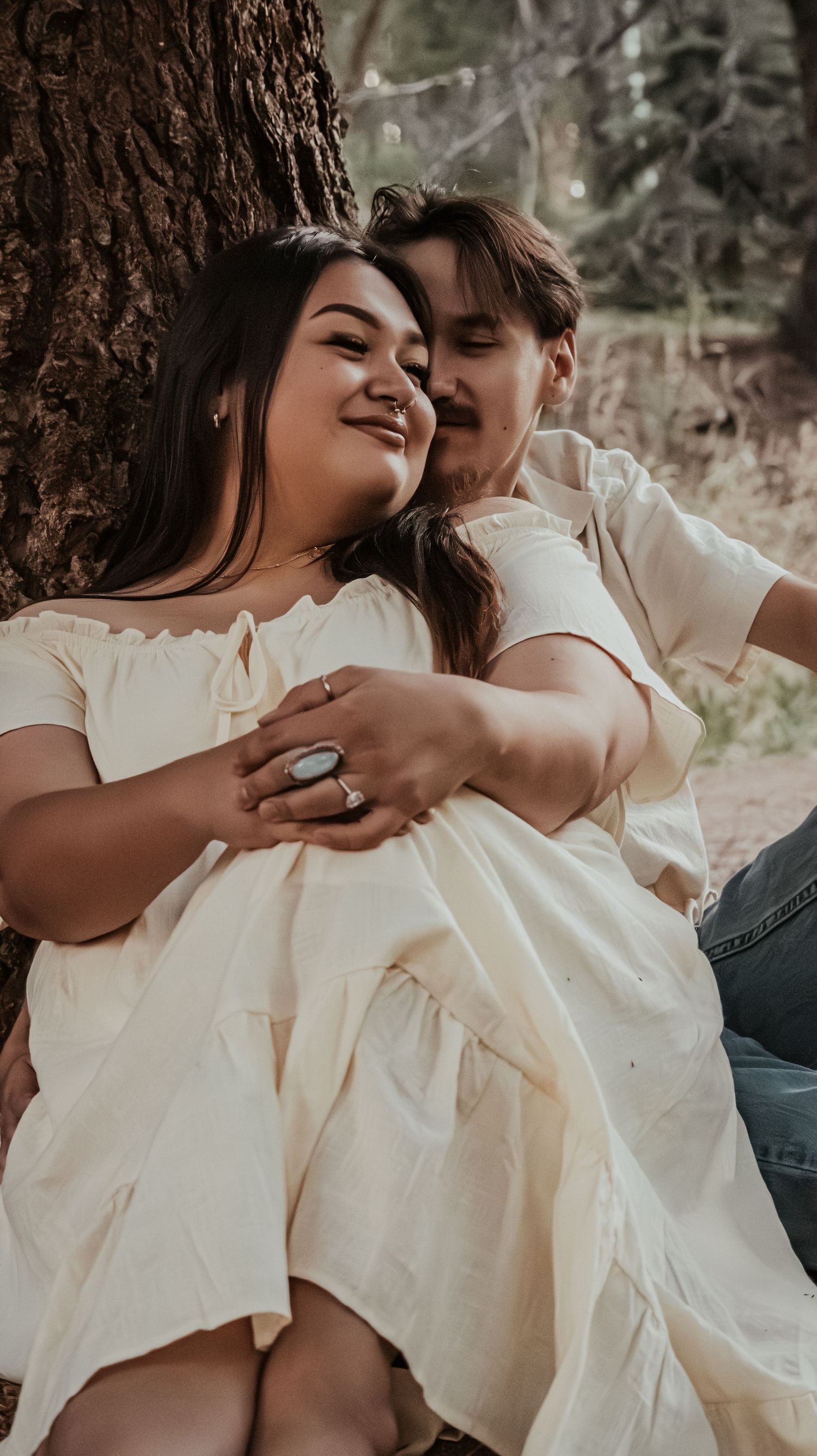 A couple sits against a tree, the woman in a cream dress smiling as her partner embraces her warmly.