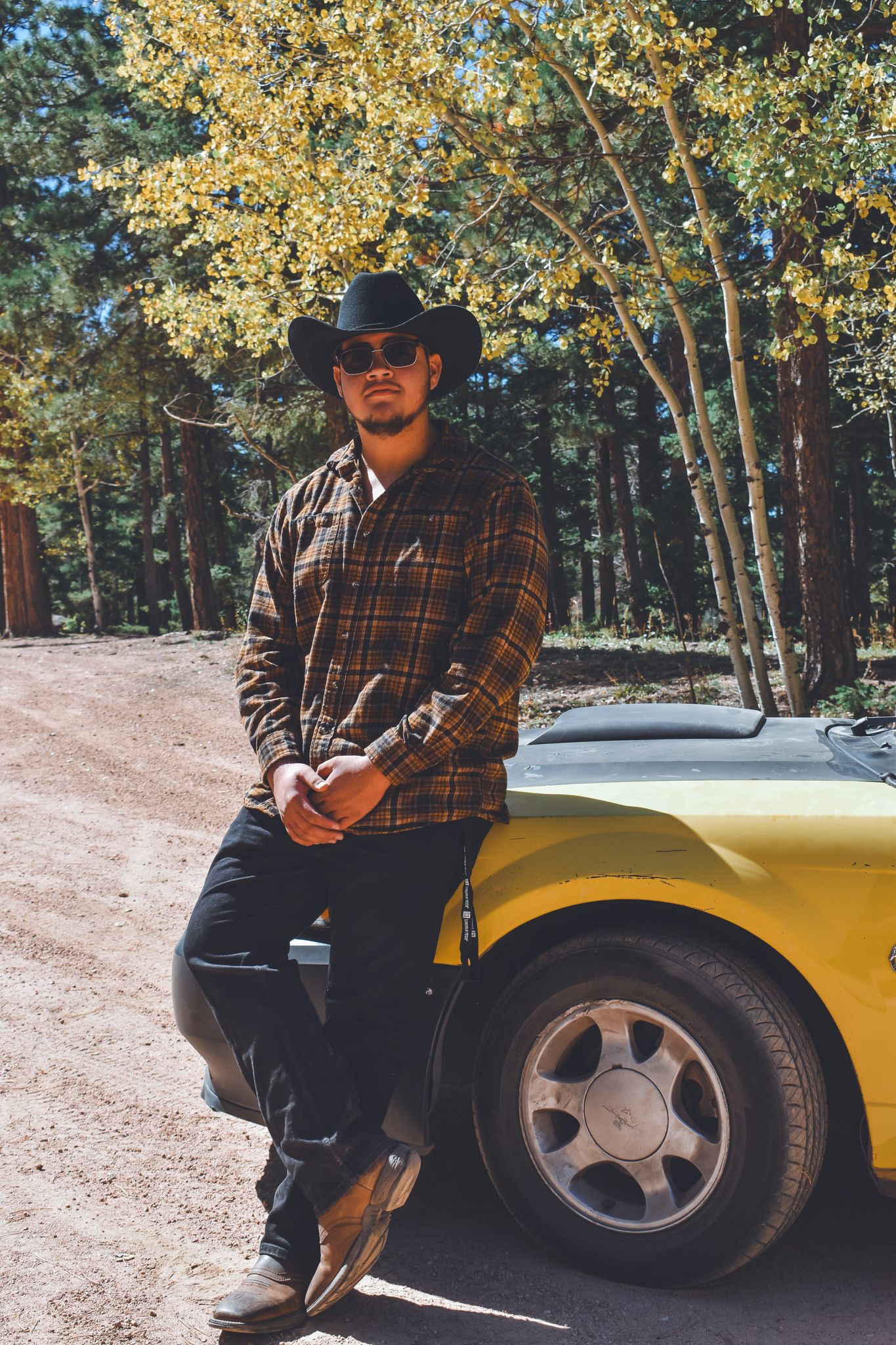 Man in cowboy hat and plaid shirt leaning against a yellow sports car in a forest for senior photos.