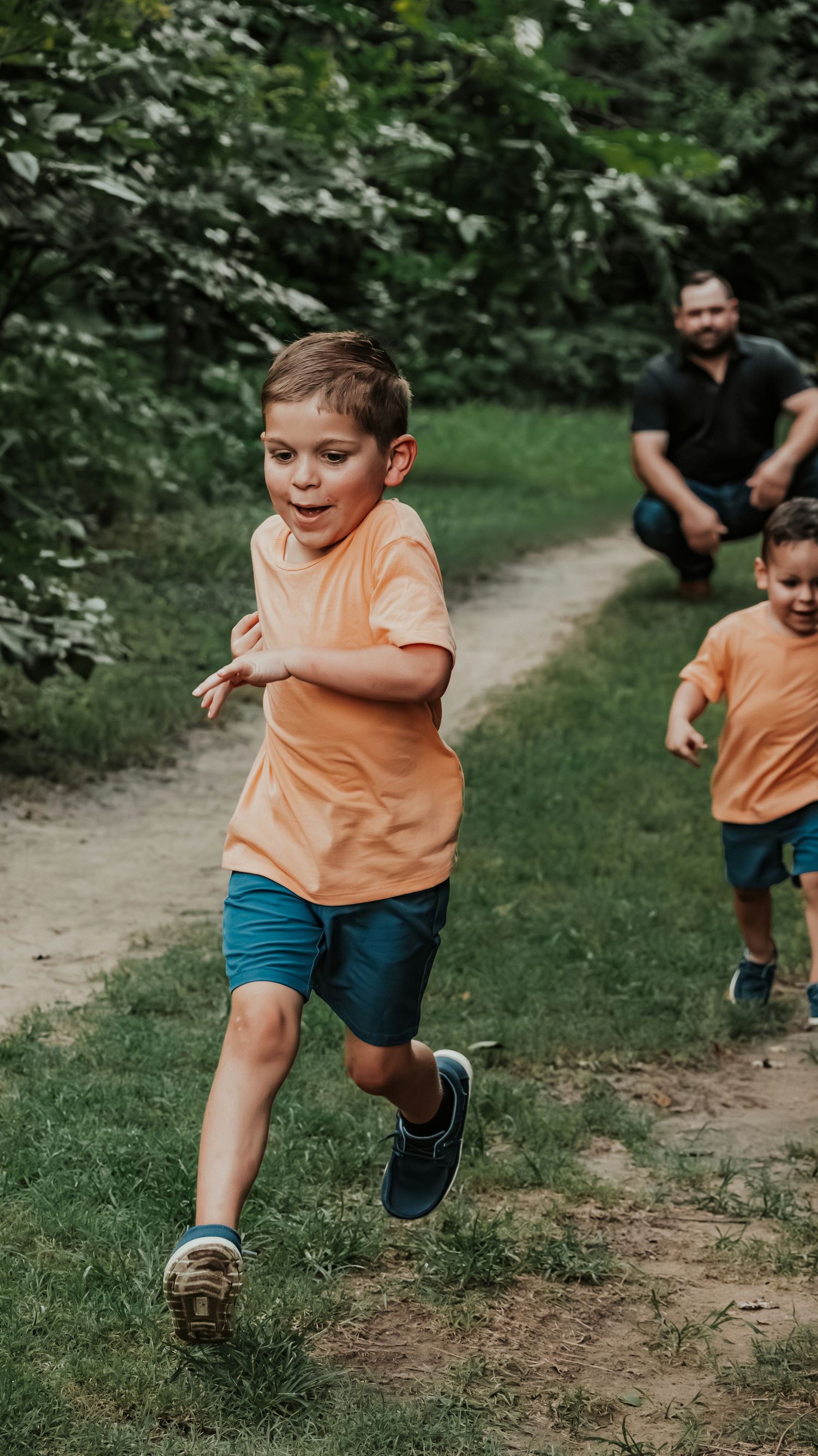 Two young boys in matching orange shirts and blue shorts racing down a wooded path with dad behind them.