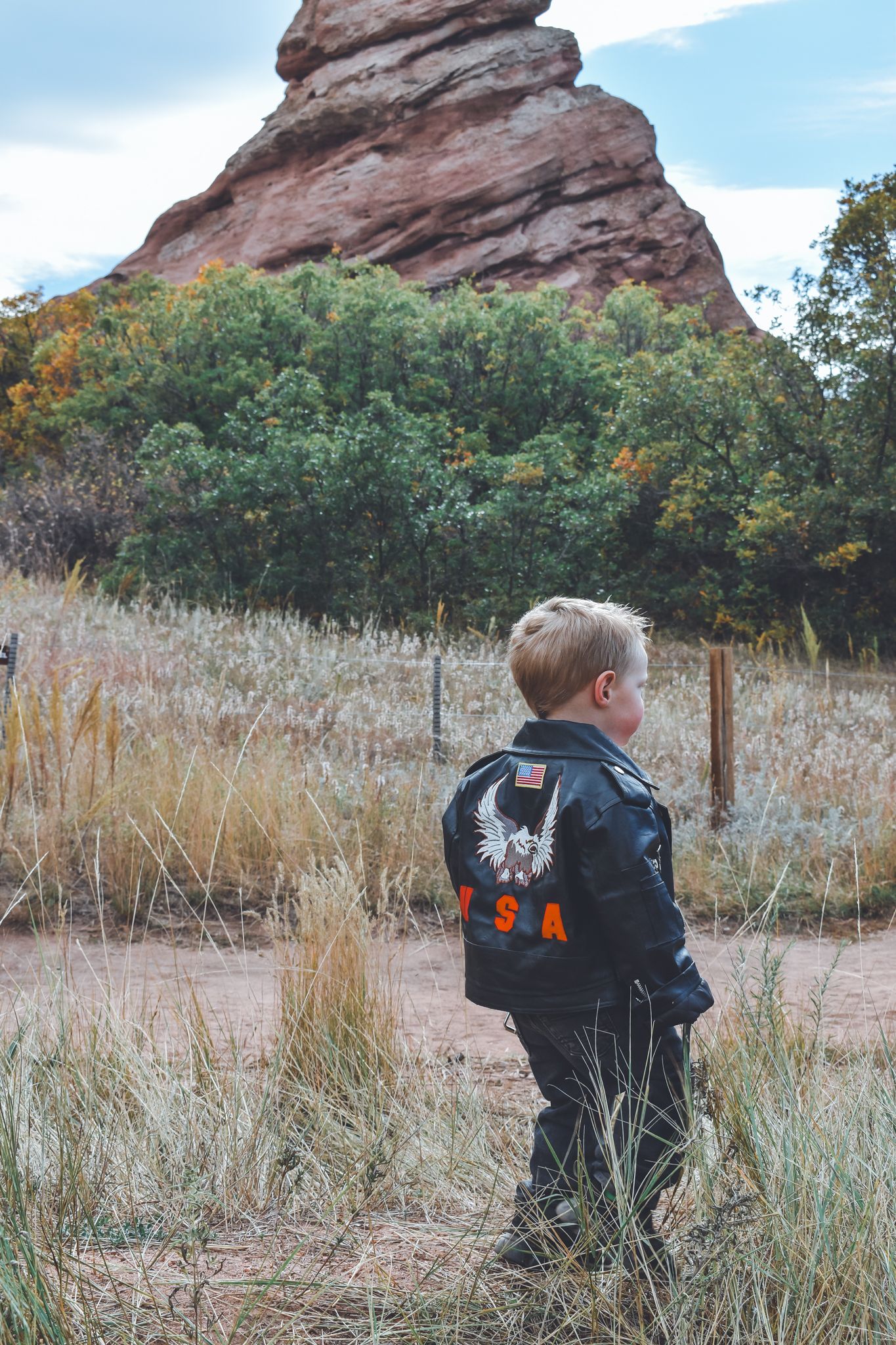 Young boy wearing USA biker jacket standing in field with red rock formation behind him. Family Photographer in Pflugerville, Tx