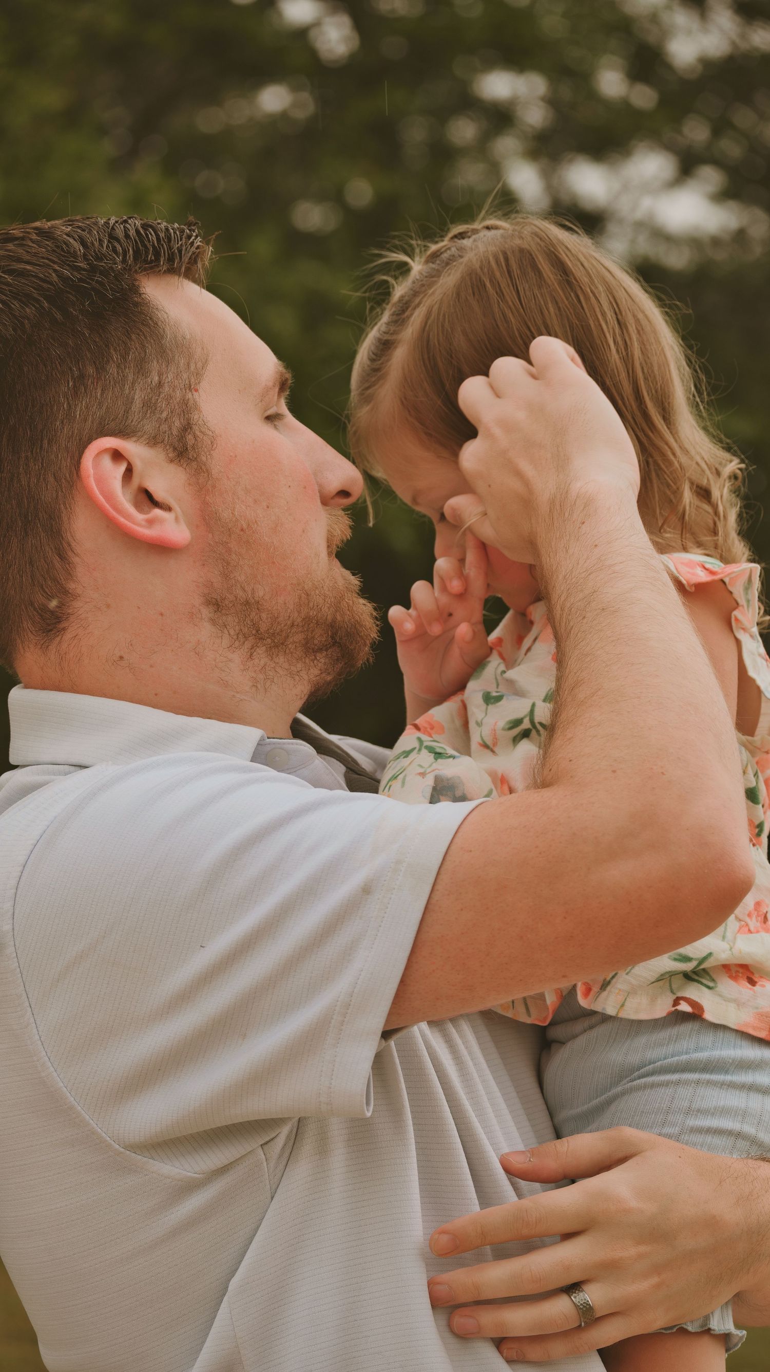 A father holds his young daughter outdoors, gently touching her forehead in a loving moment for a family session.
