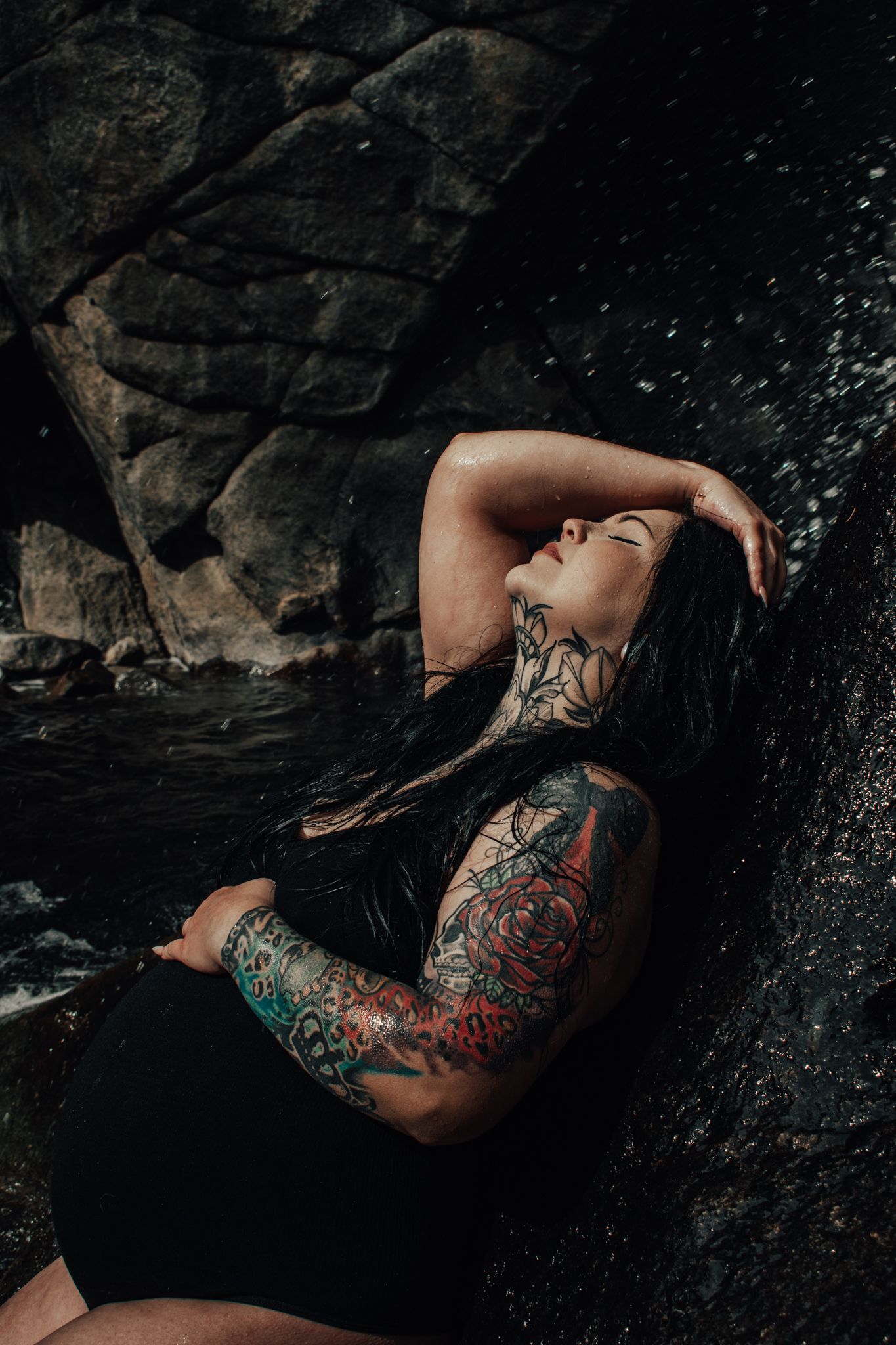 Tattooed woman in black outfit leans back against dark rocks near a waterfall. Maternity Photographer in Pflugerville, Tx