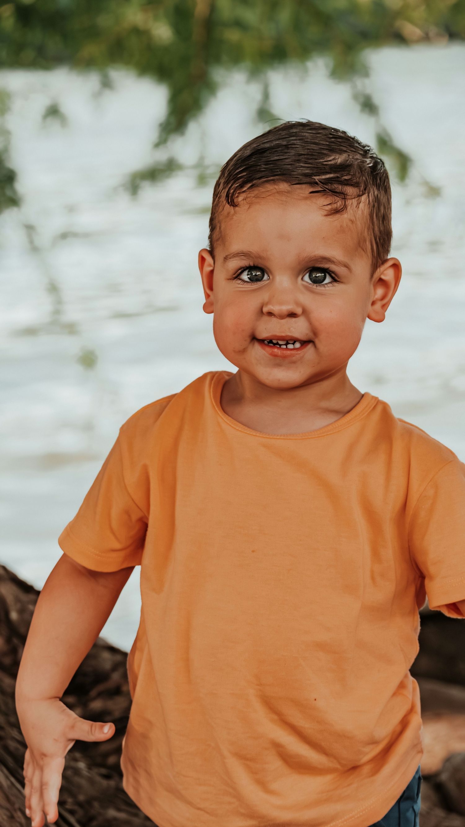 Adorable toddler boy in an orange t-shirt smiling at the camera near a sunlit water background.