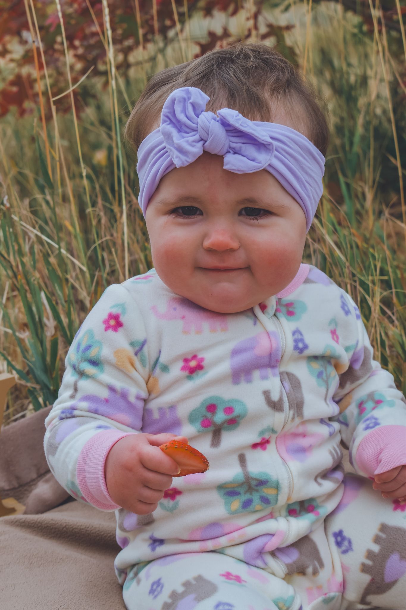 Adorable baby girl and floral onesie sitting outdoors holding a small snack. Family Photographer in Pflugerville, Tx