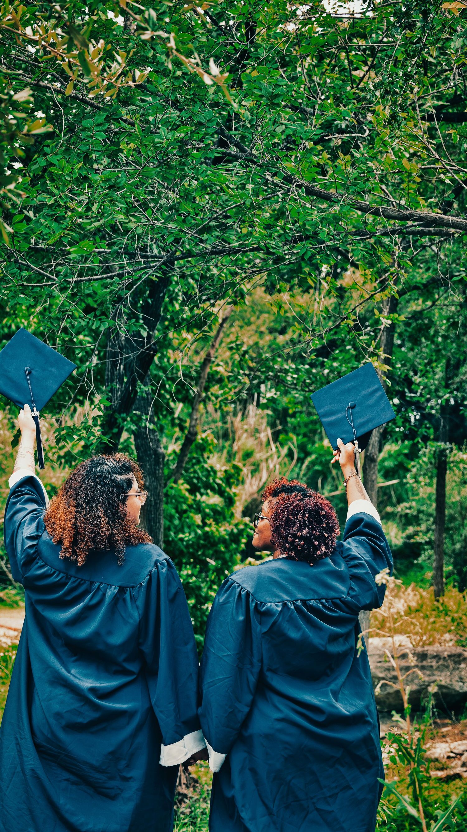 Two female graduates in blue robes hold caps up beneath green trees in nature for senior photos.