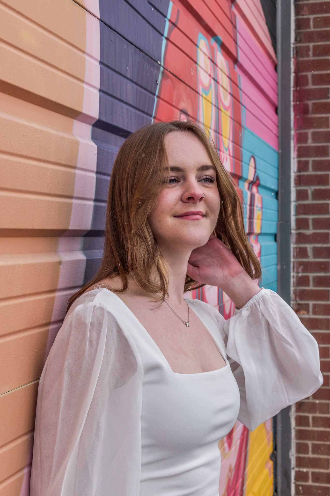Young woman in white square-neck top smiling softly while leaning against vibrant colorful graffiti wall for senior photos.