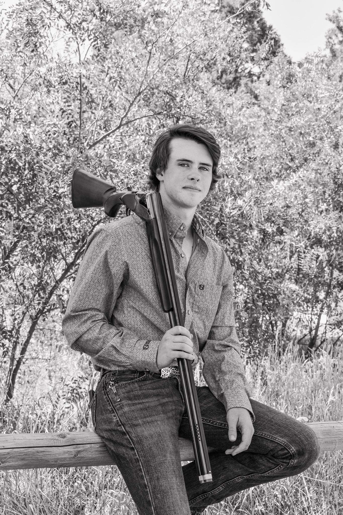 Black and white portrait of young man holding a shotgun while leaning on a wooden fence outdoors for senior photos.