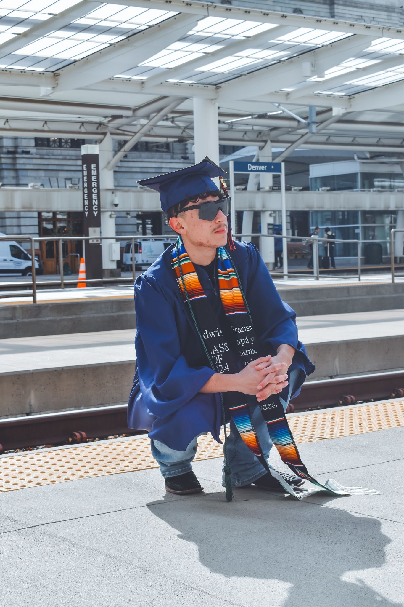 Graduate in blue cap and gown crouches on Denver train platform wearing sunglasses and colorful stole for senior photos.