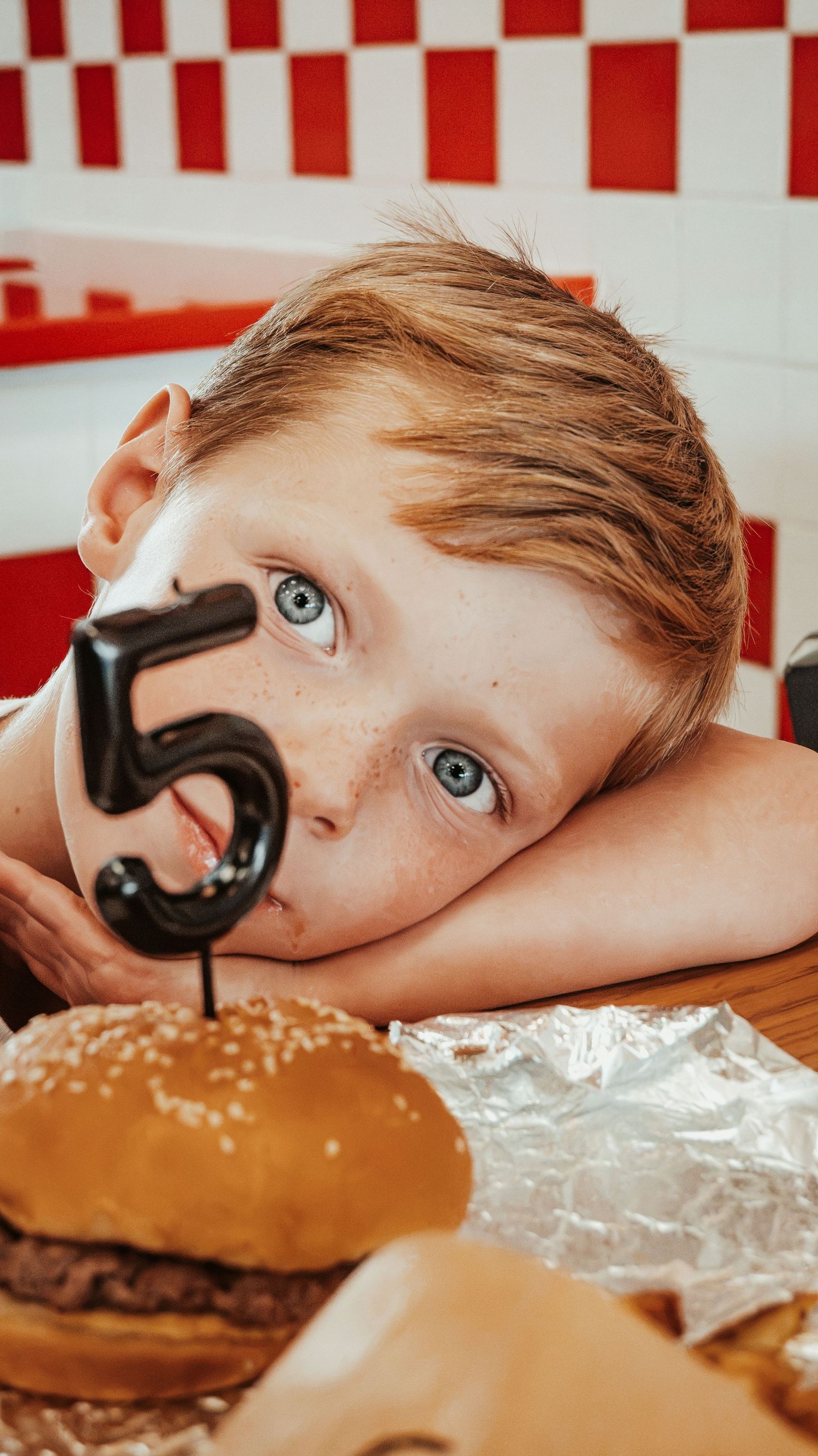 Blue-eyed boy resting chin on table next to a burger with number 5 candle at Five Guys.
